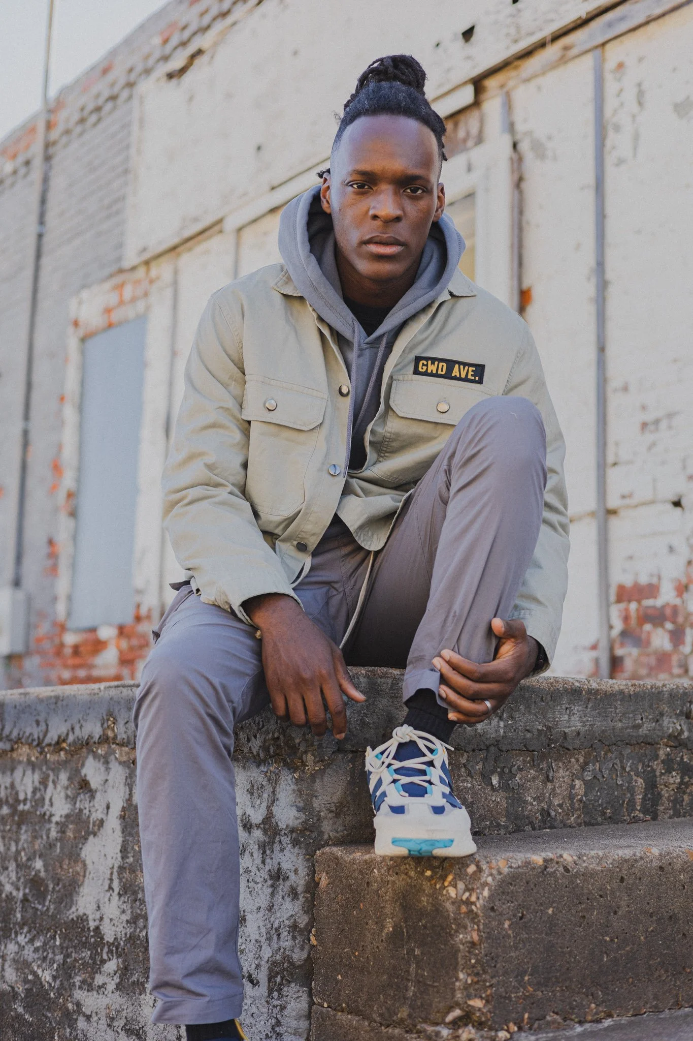 A young Black man with dreadlocks tied up in a bun, wearing a beige jacket with a GWD AVE patch, a gray hoodie, gray pants, and sneakers, sitting on outdoor concrete steps in front of a weathered brick wall with some paint peeling off.