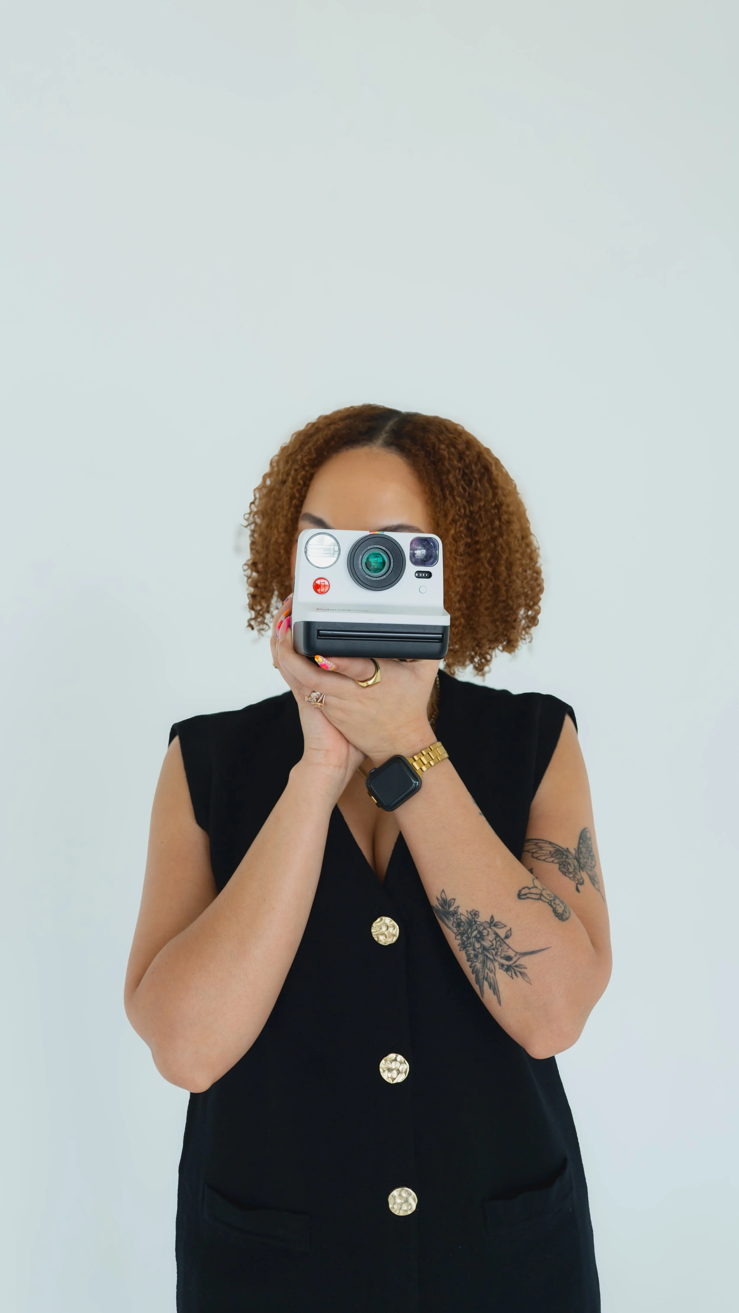 A woman with curly, shoulder-length hair wearing a black sleeveless dress with gold buttons, holding a vintage instant camera to her face, revealing tattoos on her arms, a gold watch, ring, and bracelet, against a plain white background.