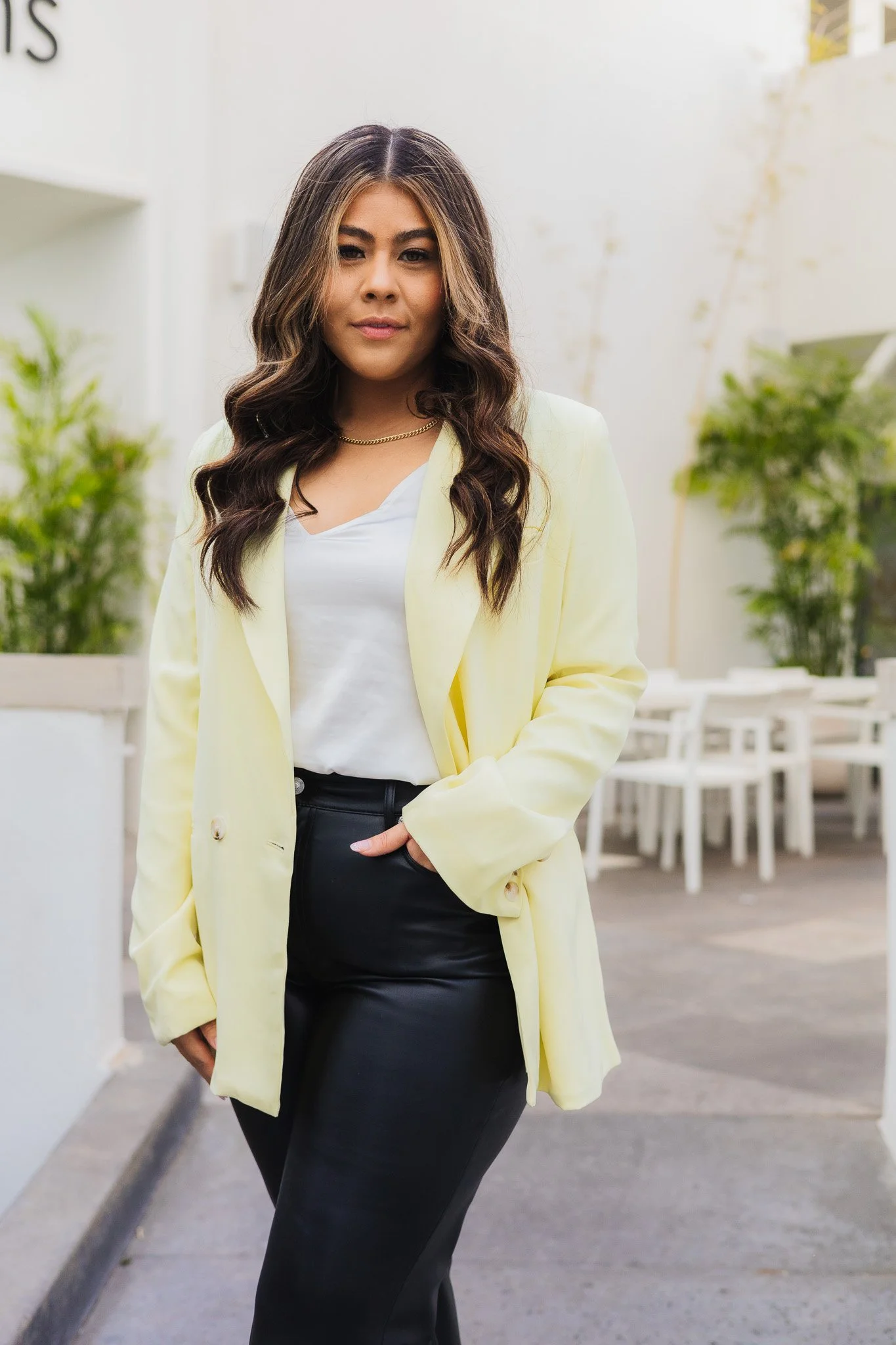 A woman with long wavy dark hair wearing a light yellow coat, white top, black leather pants, and a gold chain necklace standing outdoors with potted plants and white furniture in the background.