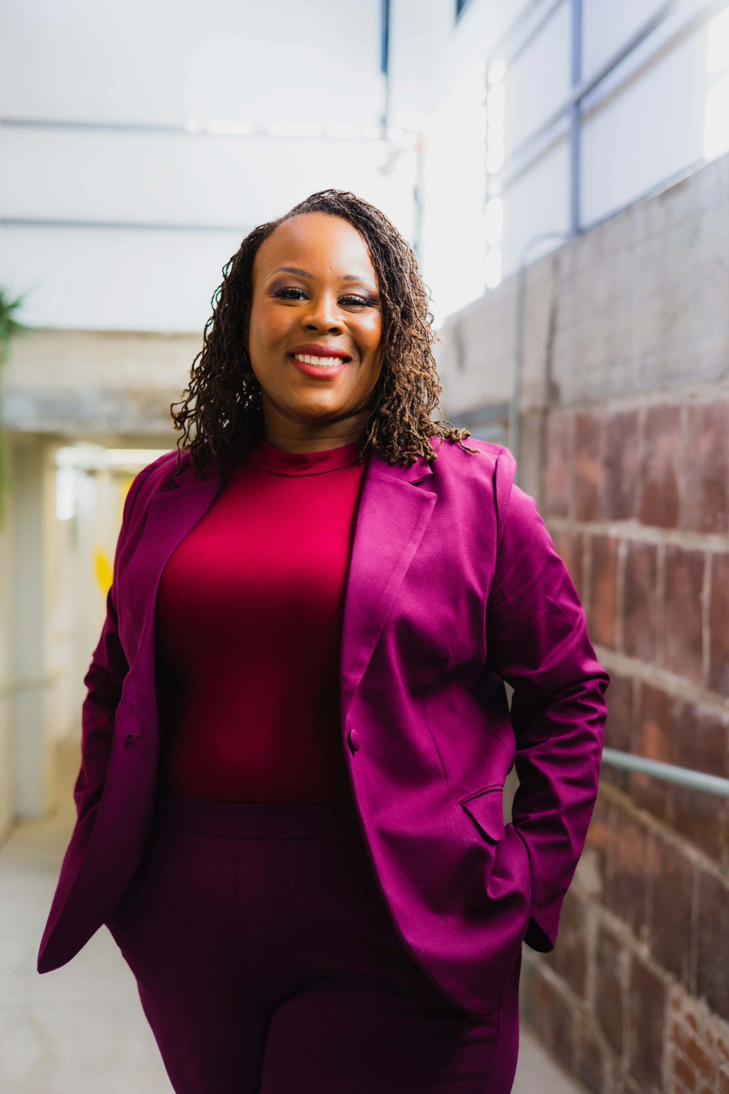 A smiling Black woman with dreadlocks wearing a magenta blazer and matching top, standing in an indoor space with brick and concrete walls and large windows.
