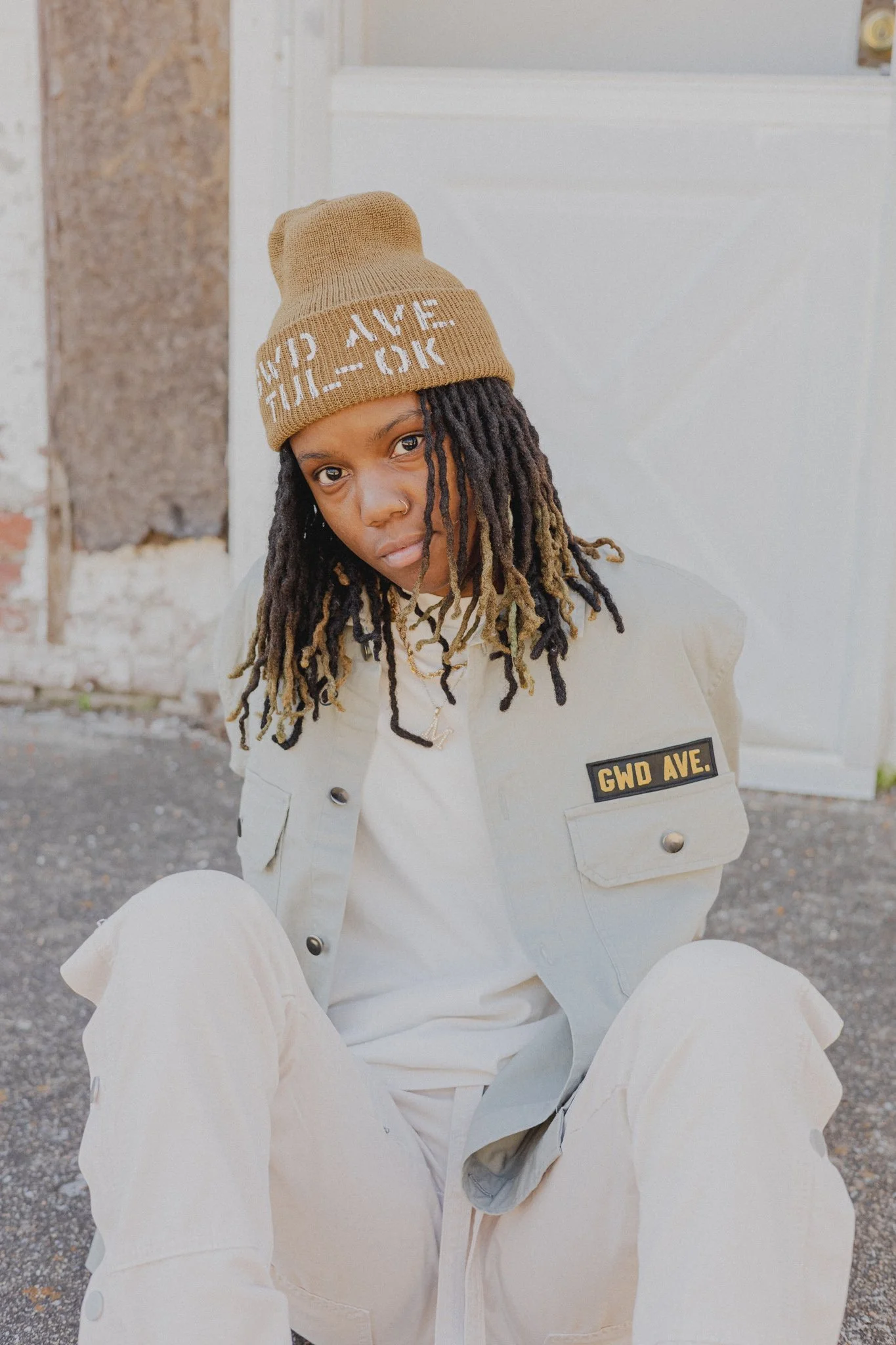 A woman with dreadlocks wearing a tan knit beanie and a beige jacket with a 'GWD AVE.' patch, sitting outdoors against a background of a partially demolished building and a white door.