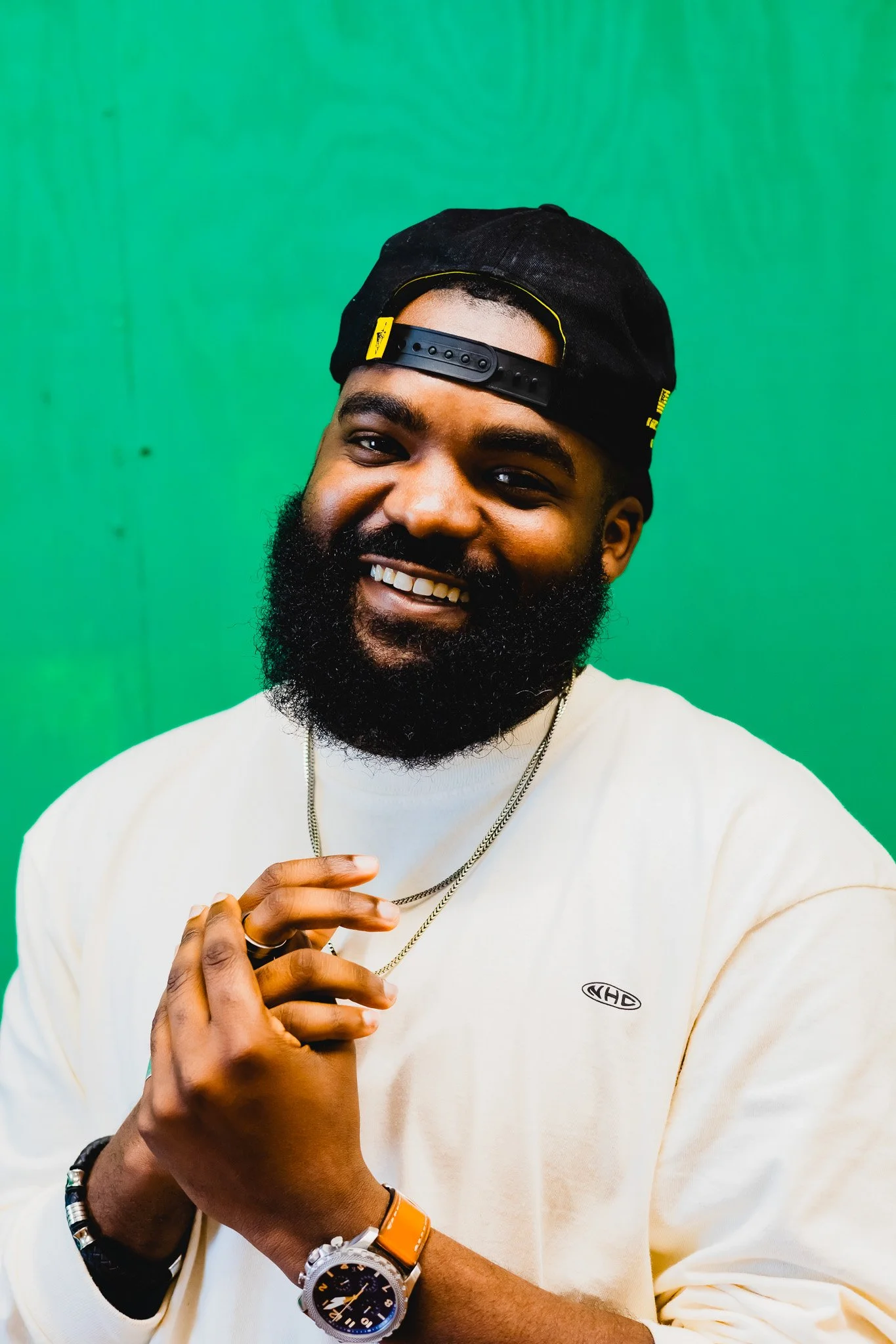 A smiling man with a beard and mustache, wearing a black baseball cap backwards, a white shirt with a small logo, and multiple accessories, including a watch, necklace, and bracelets, standing against a green background.