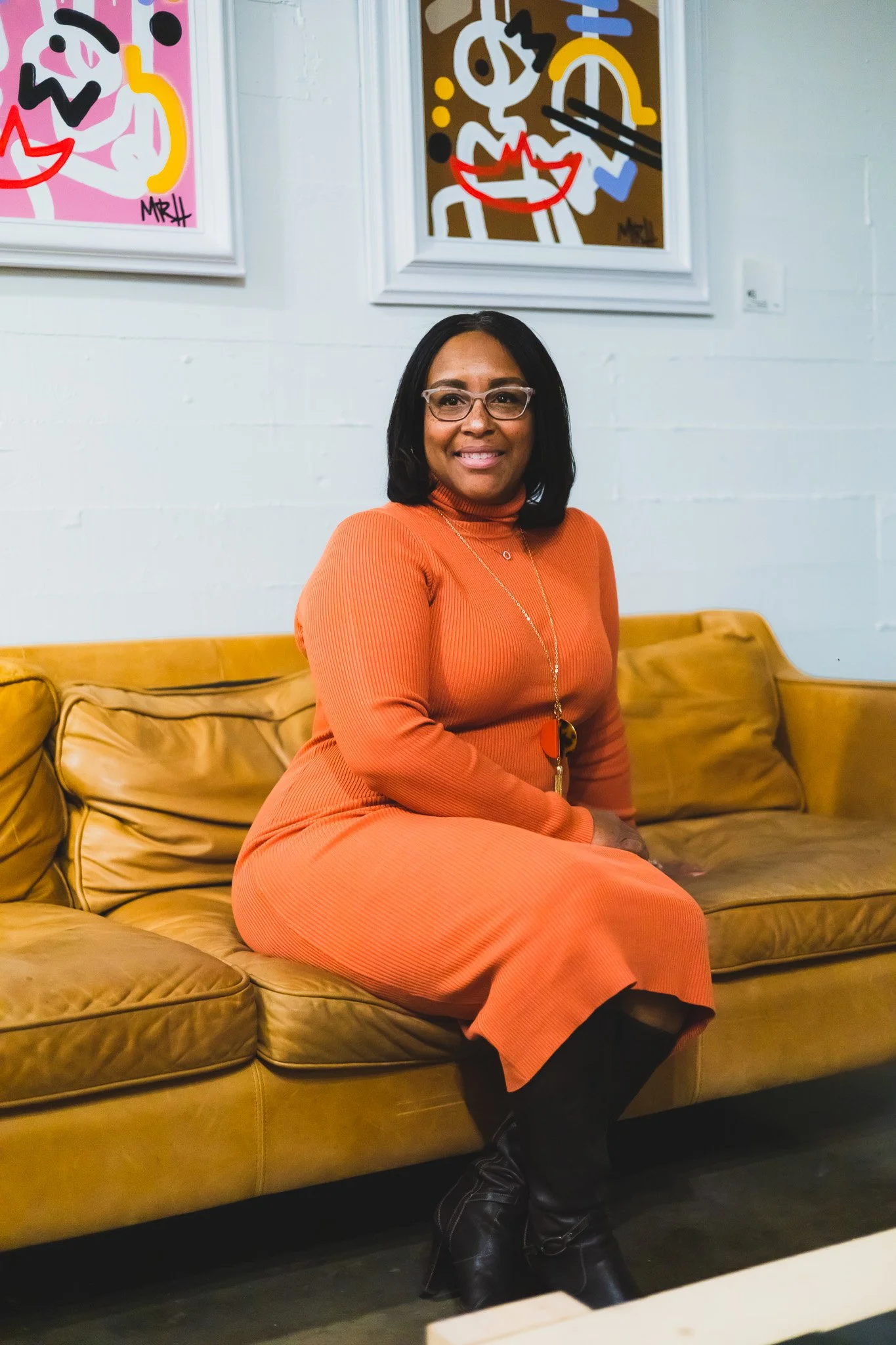 A woman with medium skin tone, black hair, and glasses, sitting on a brown leather couch in an art gallery, wearing an orange dress and black boots, smiling at the camera. There are two colorful abstract paintings on the white wall behind her.