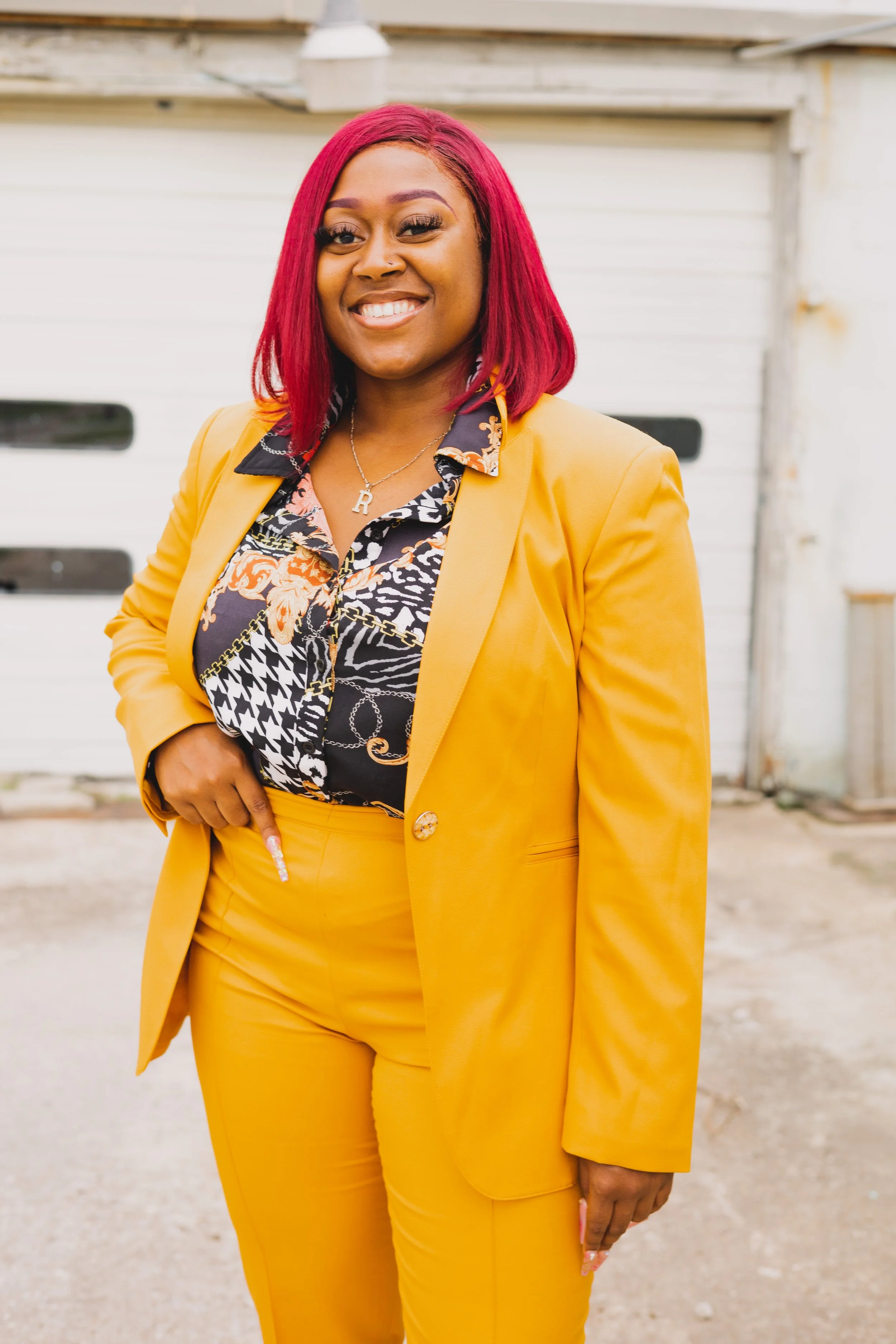 A woman with bright red hair smiling in front of a white garage door, wearing a yellow suit with a patterned shirt underneath, and a necklace with the letter 'R'.
