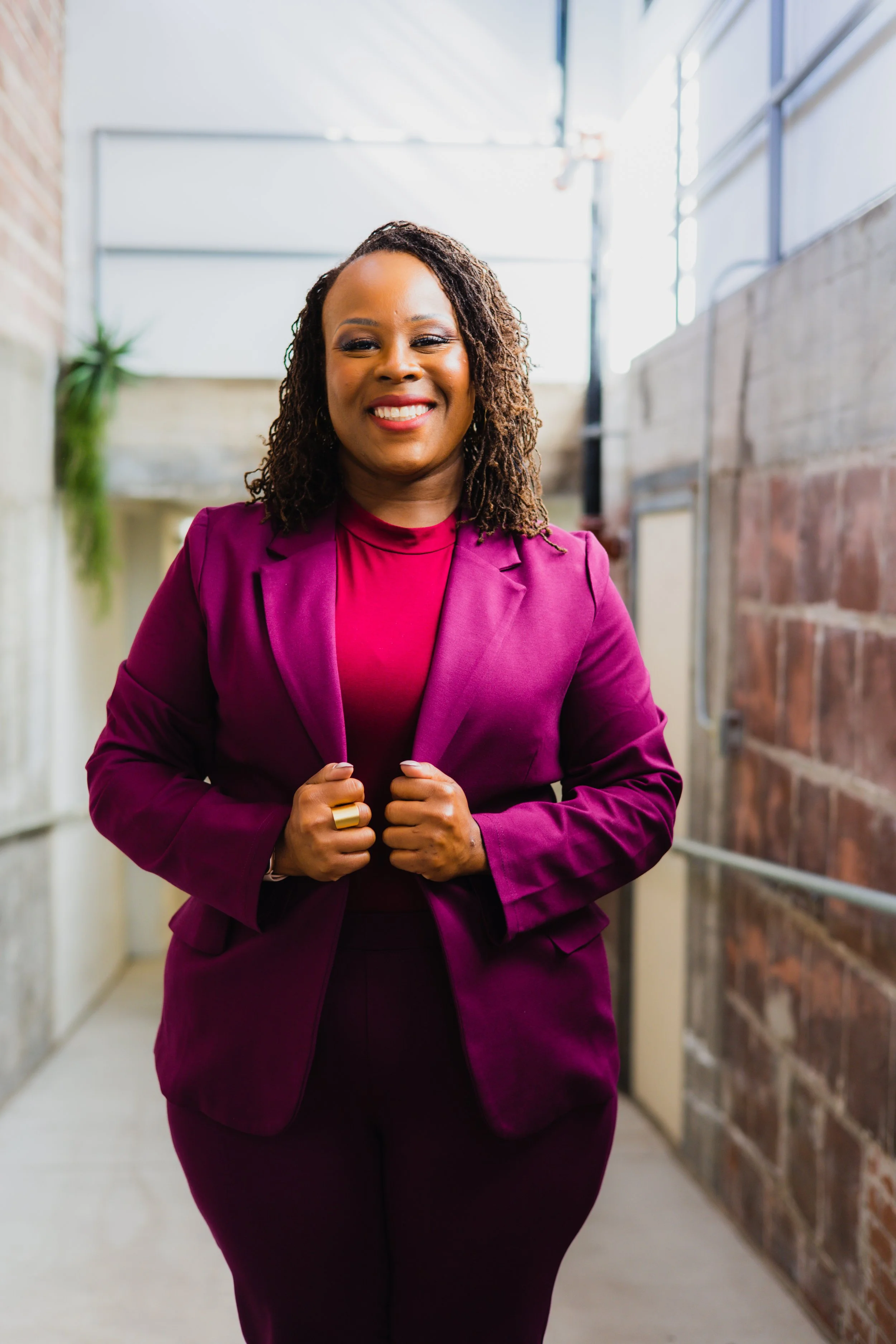 A confident Black woman in a magenta blazer and matching pants standing in an industrial setting with brick and concrete walls, smiling at the camera.