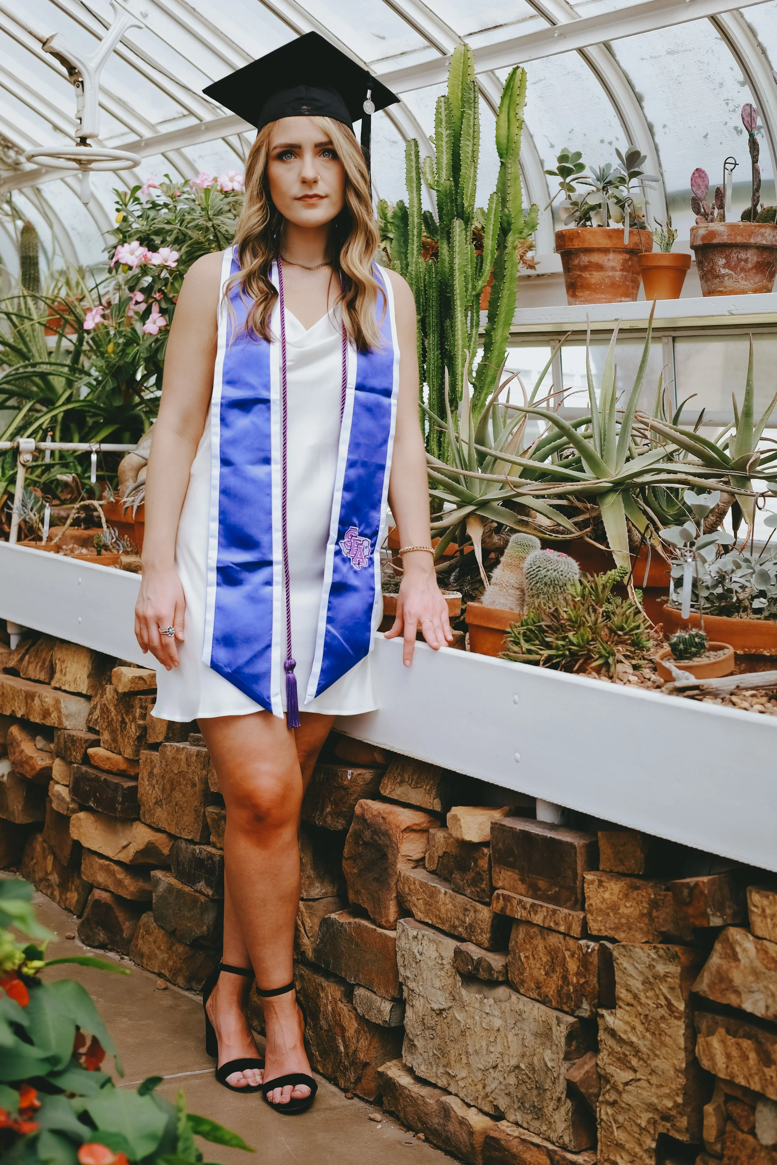 A woman wearing a graduation cap and gown in a greenhouse surrounded by various cacti and succulents.