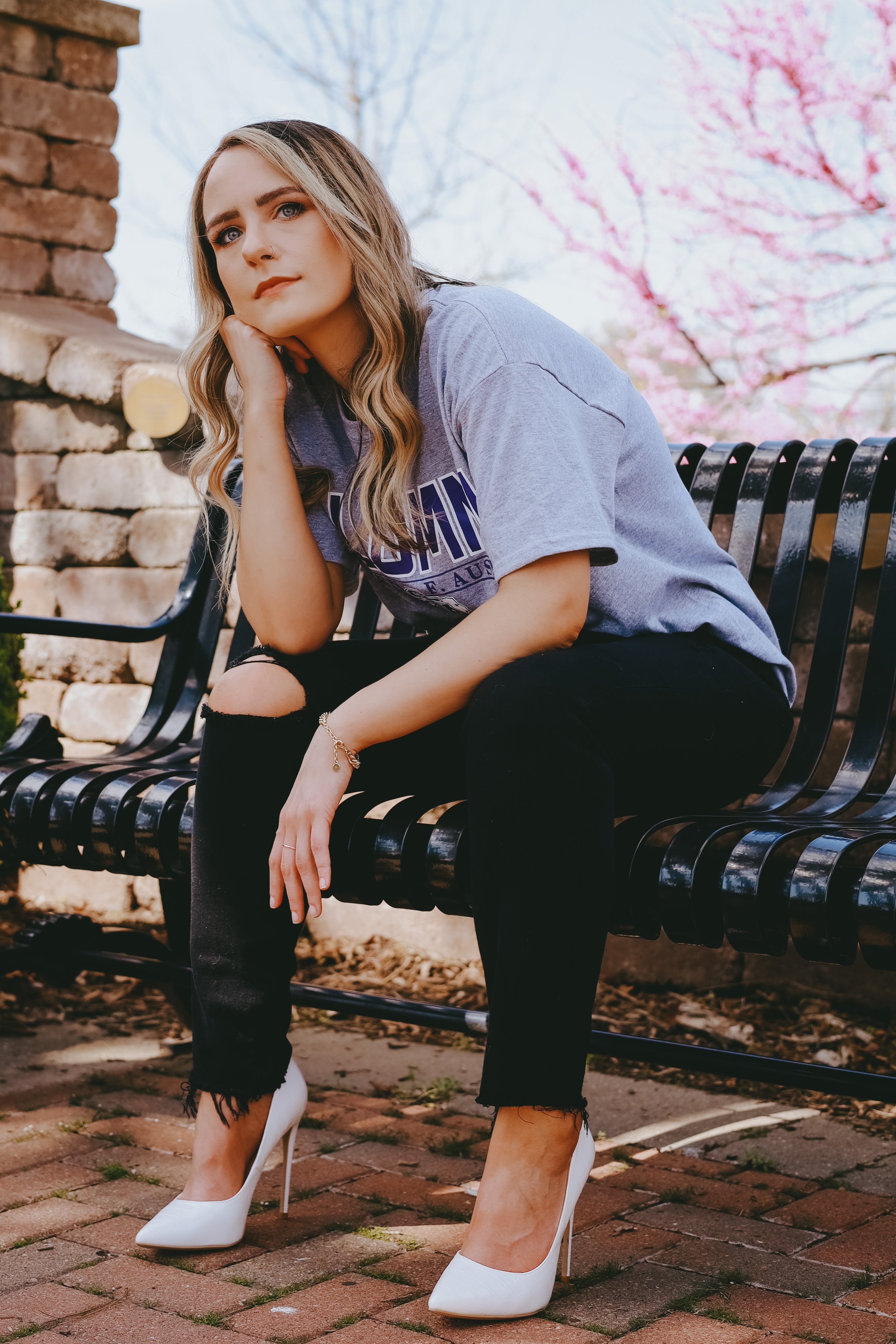 A young woman with long wavy blonde hair, light makeup, wearing a loose gray T-shirt, ripped black jeans, and white high heels, sitting on a black metal park bench with her head resting on her hand. Behind her, pink cherry blossom trees are in bloom,