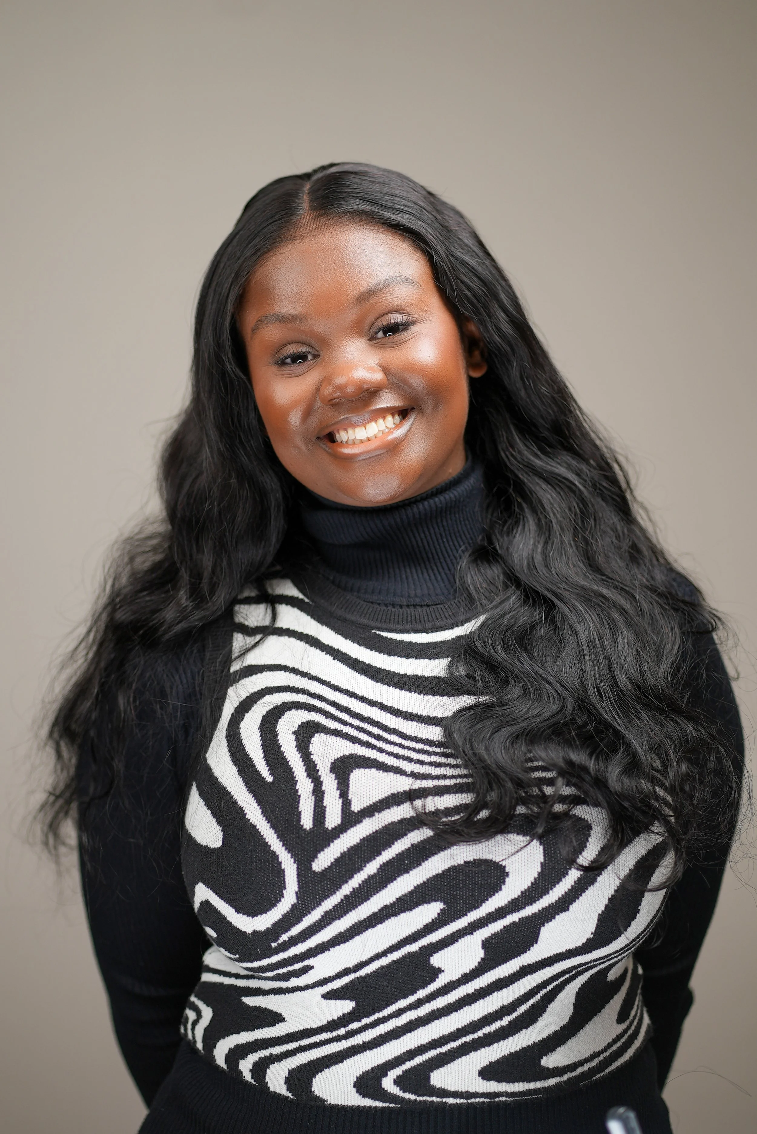 A smiling African American woman with long, black, wavy hair, wearing a black and white patterned sweater and a black turtleneck, standing against a plain beige background.
