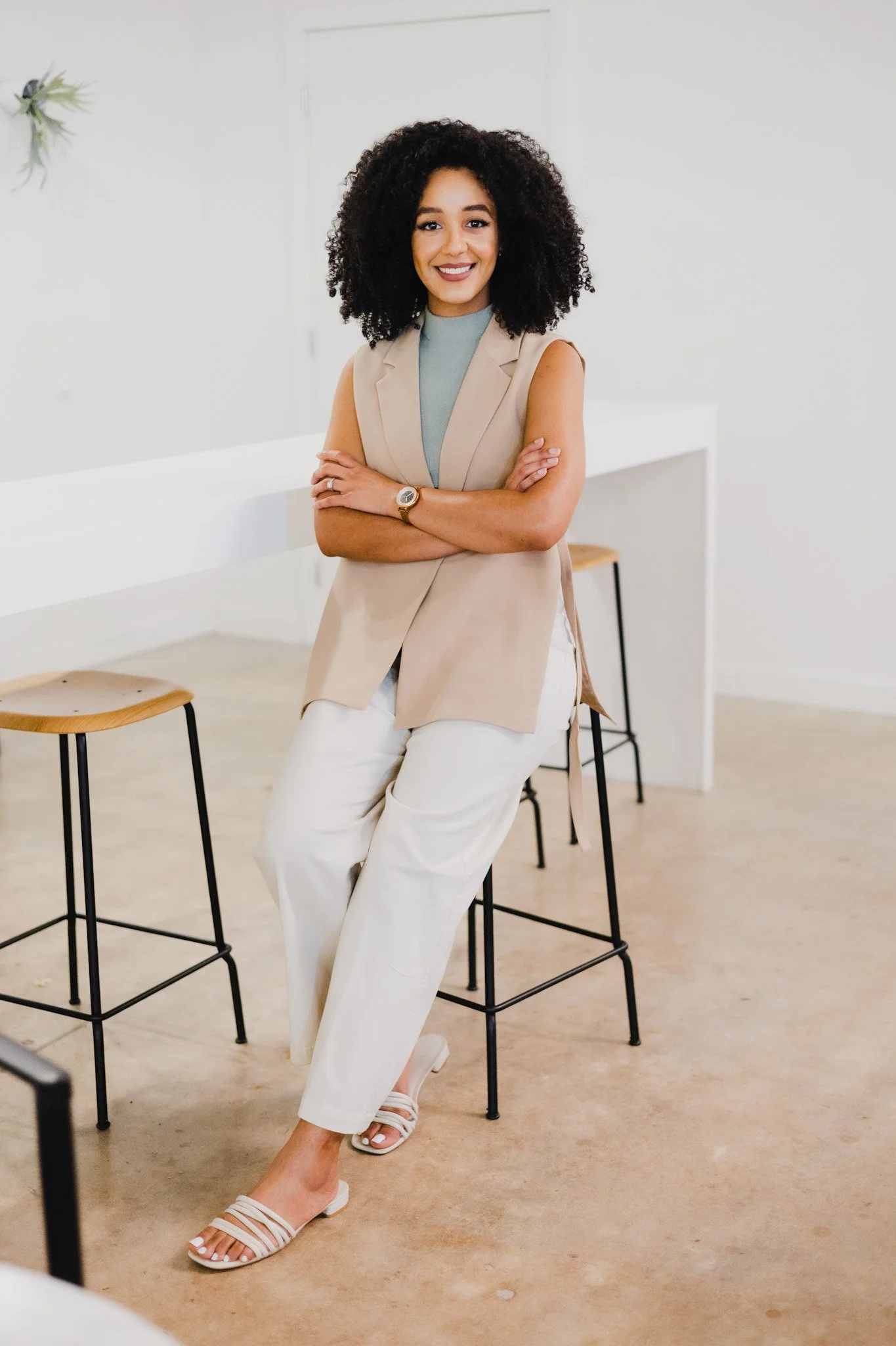 A woman with dark curly hair, smiling, wearing a beige sleeveless blazer, light blue top, white pants, beige sandals, sitting on a stool with crossed arms in a modern, minimally decorated room with white walls and wooden stools.