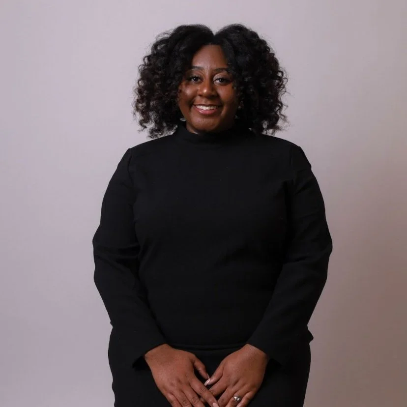 A woman with curly black hair wearing a black top, standing against a plain light-colored background, smiling at the camera.