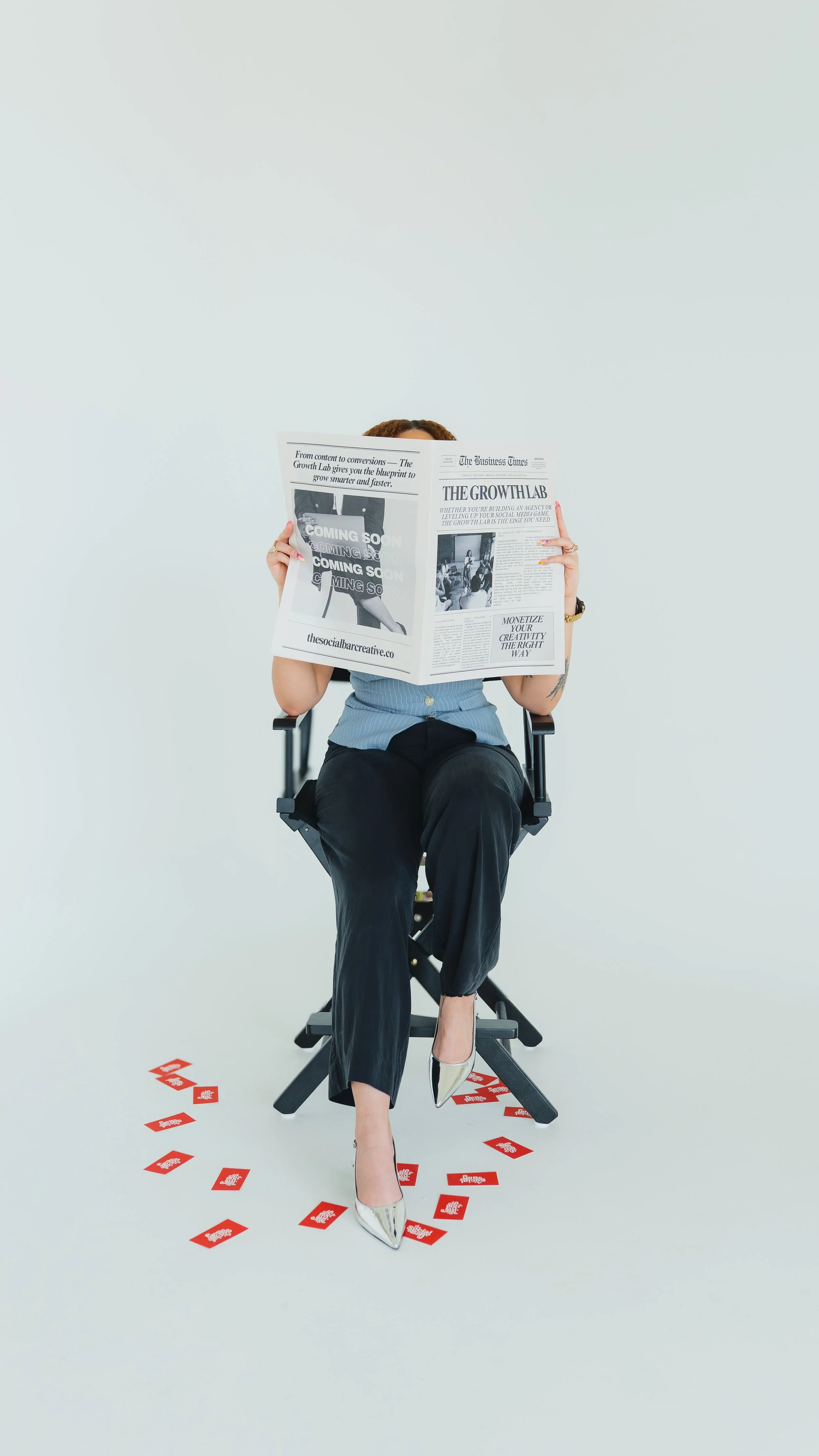 A woman sitting on a director's chair, reading a newspaper, with scattered red and white cards on the floor around her.