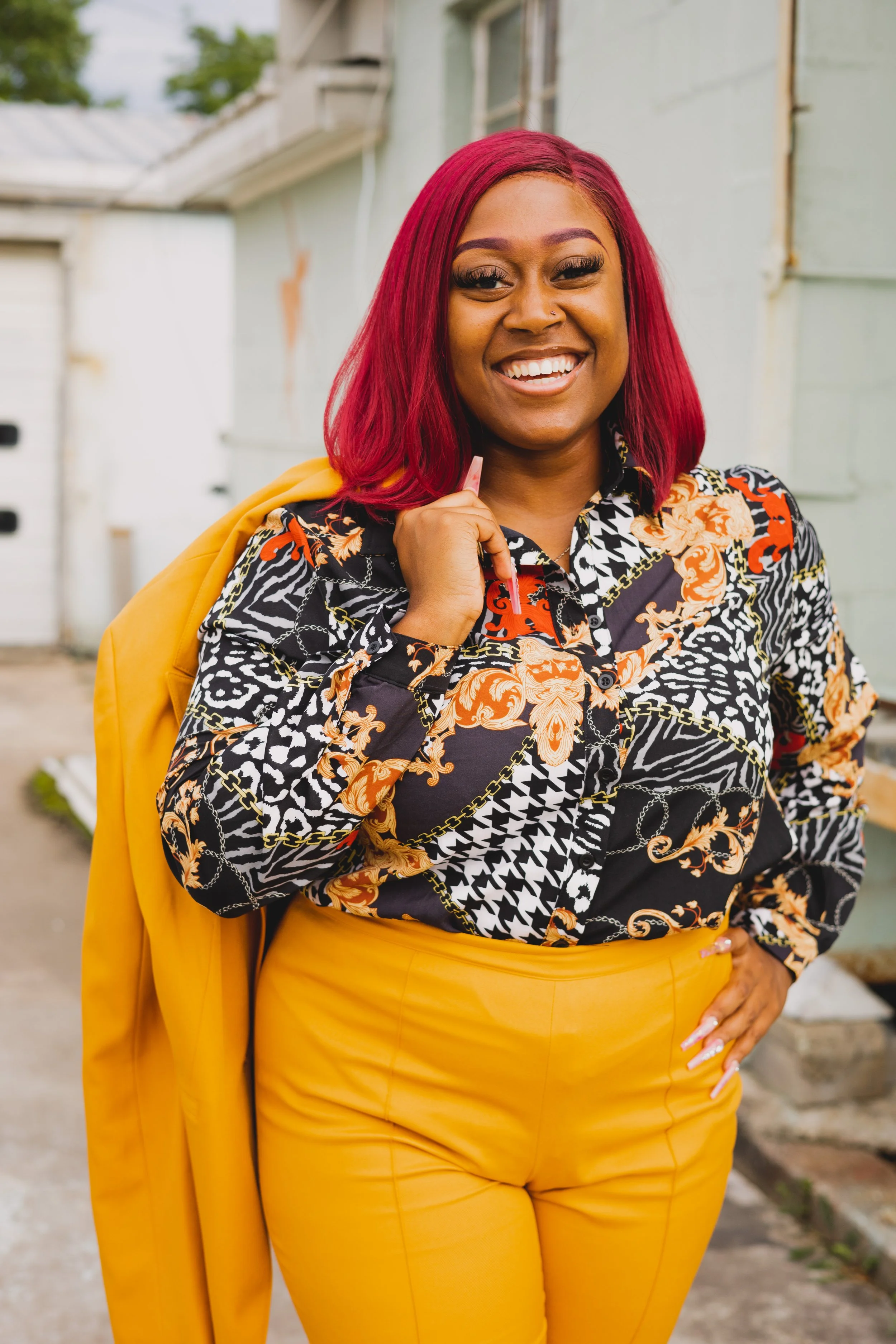 A woman with bright red hair, smiling and holding a straw, wearing a colorful patterned blouse and yellow pants, standing outdoors with a gray building in the background.
