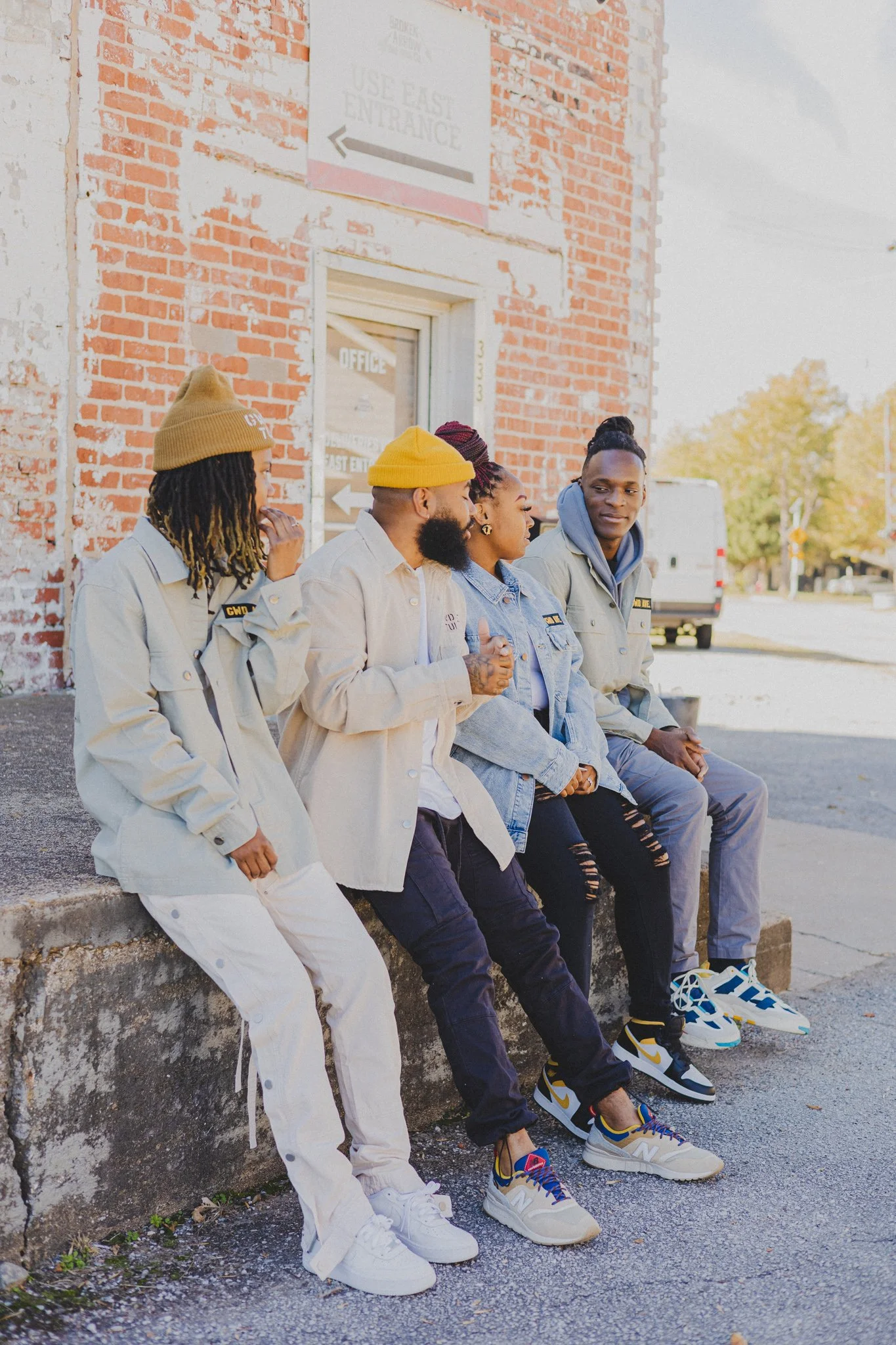Four people sitting on a sidewalk outside a brick building, wearing casual clothing and sneakers, with a vehicle and trees in the background.