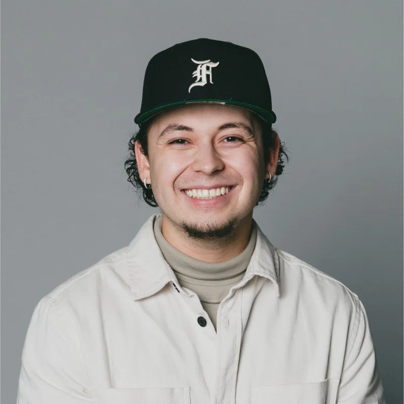 Smiling man wearing a black baseball cap with a white logo, earrings, a beige jacket over a turtleneck, against a gray background.