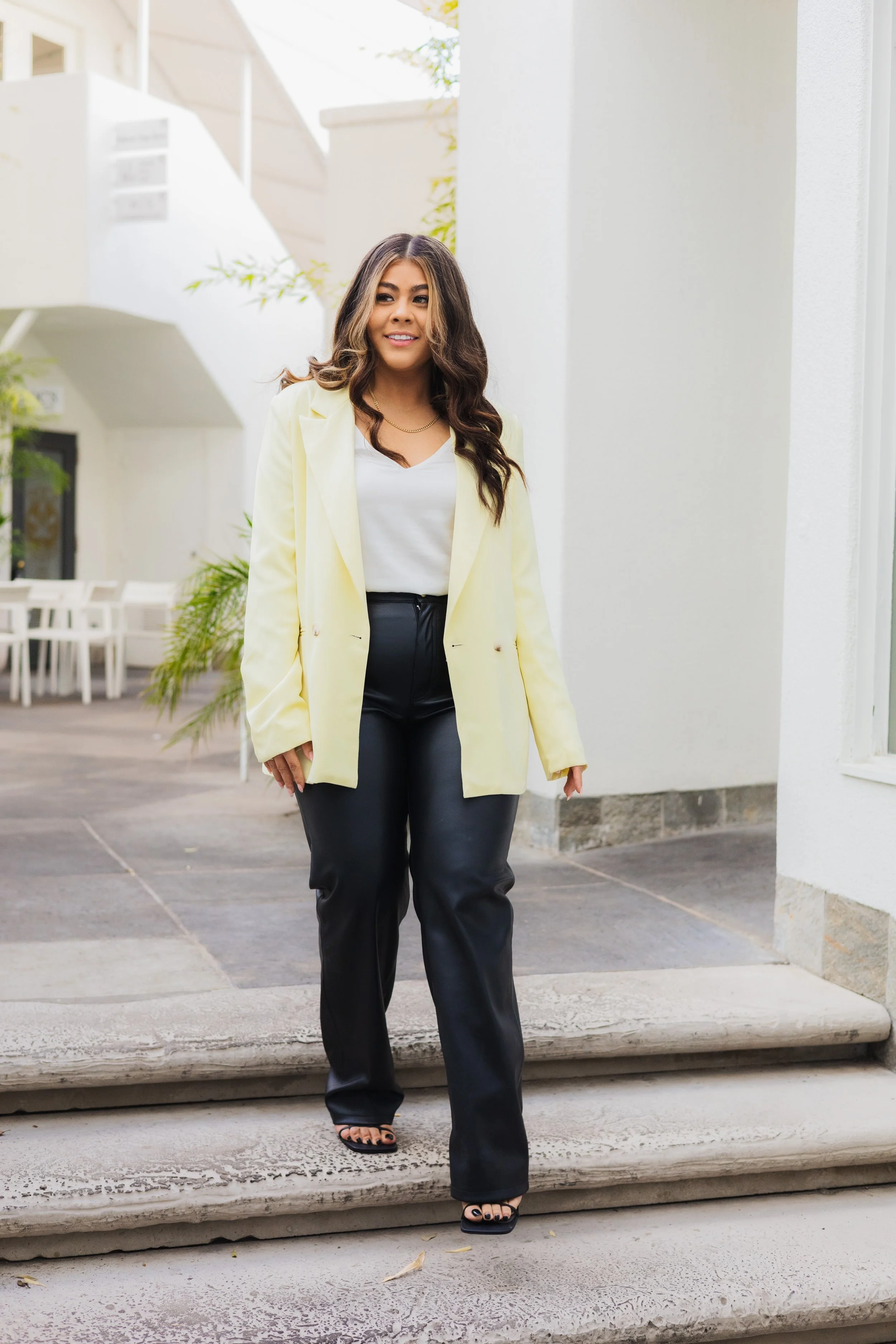 A woman walking down concrete steps outside a white building, wearing a yellow blazer, white top, black leather pants, and black strappy sandals.