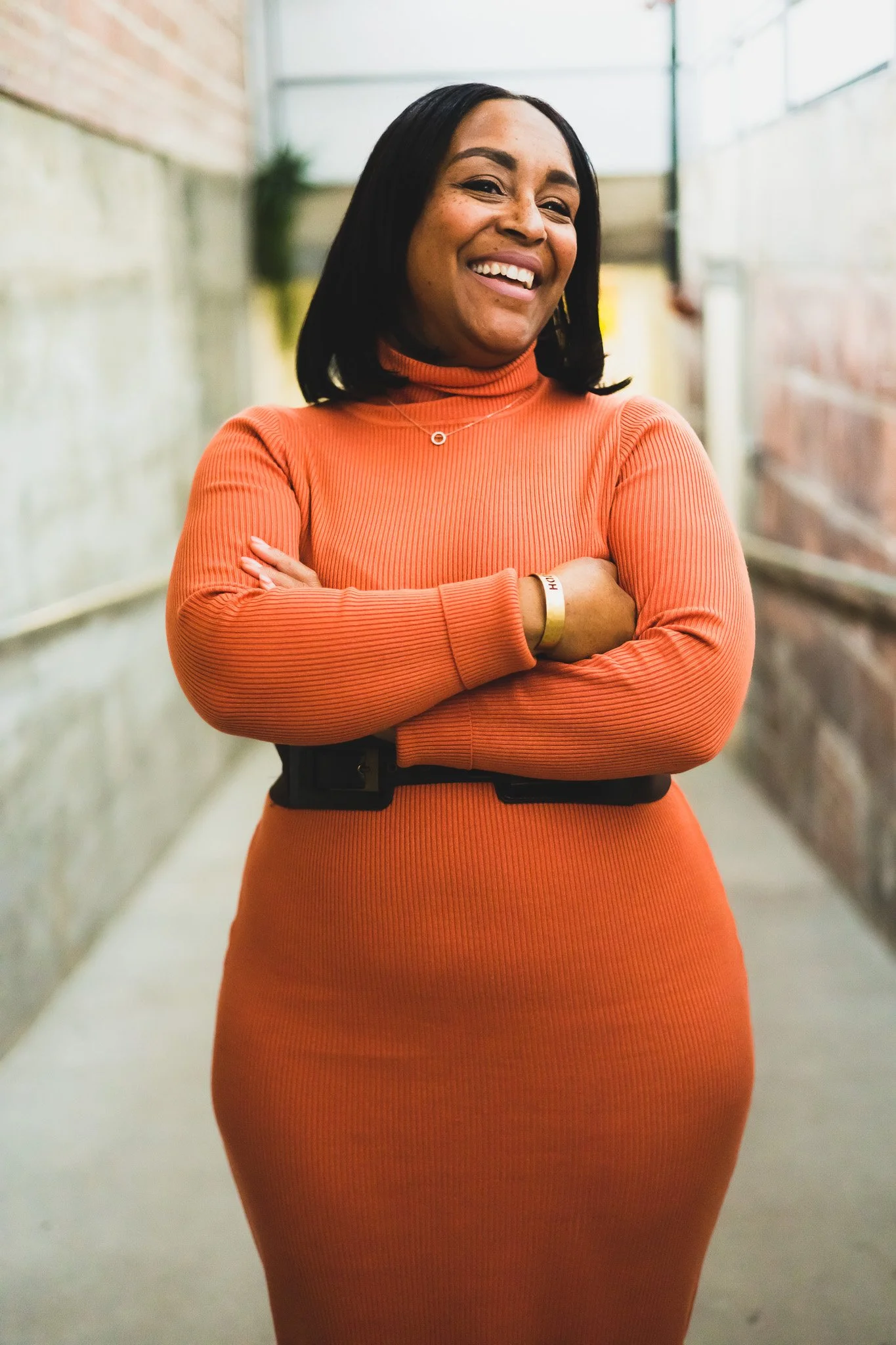 A woman with shoulder-length black hair smiling, wearing an orange turtleneck dress, standing in a brightly lit corridor with brick and concrete walls.