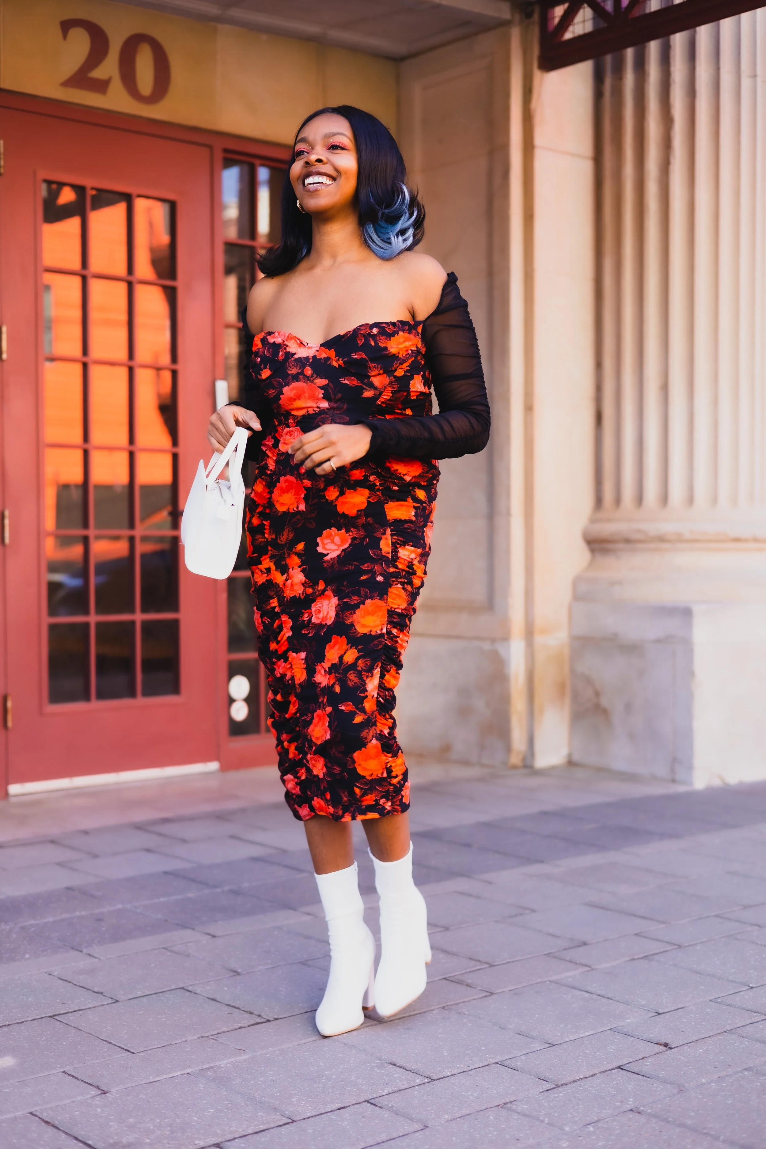 A woman in a floral dress with black and red colors, wearing white boots, is walking outside near a building with a sign reading '20' above a door. She is smiling and holding a white handbag.