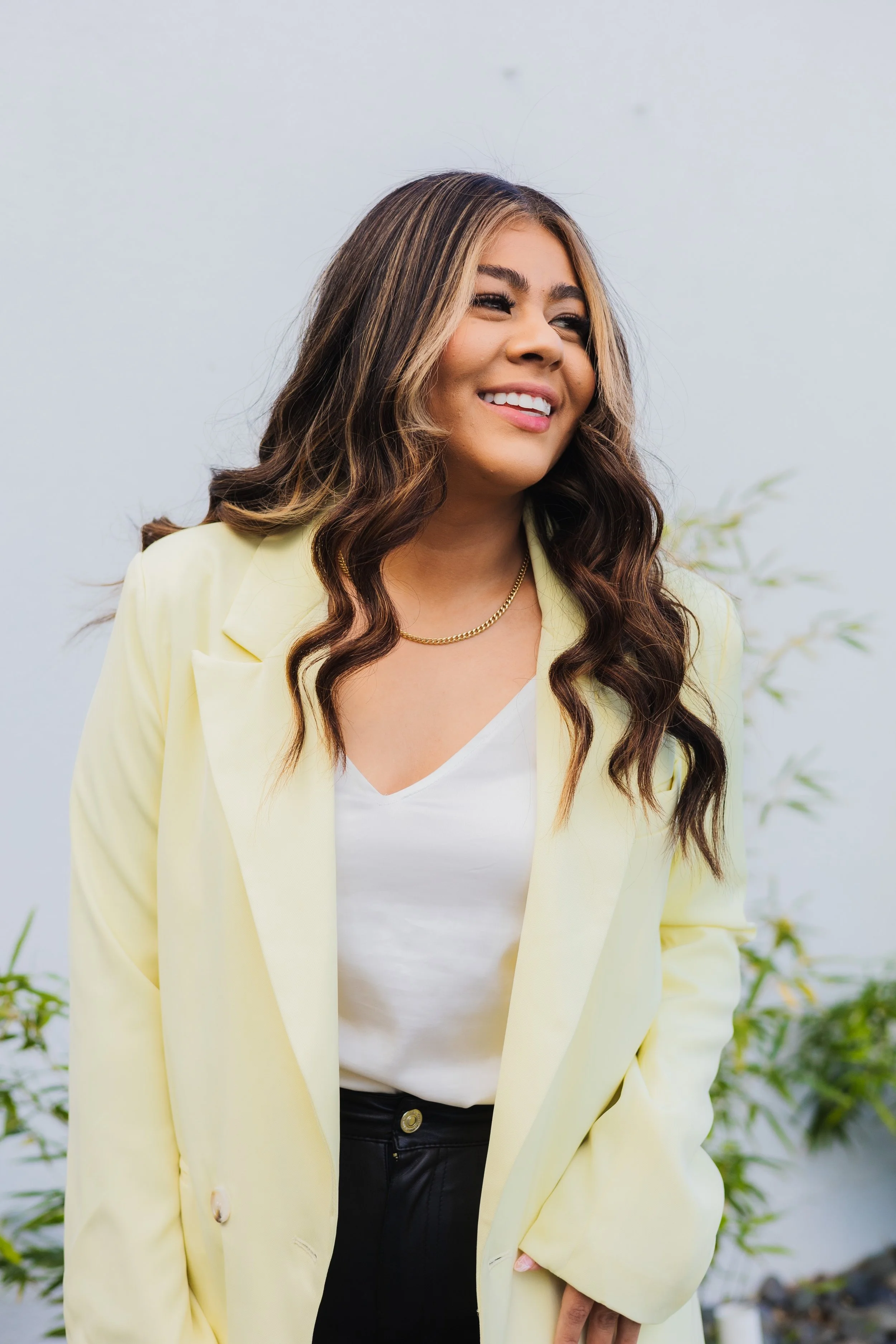 A woman with long wavy brown hair and tan skin, smiling and looking to the side, wearing a yellow blazer, white top, black pants, and gold necklace, standing outdoors with blurred greenery in the background.
