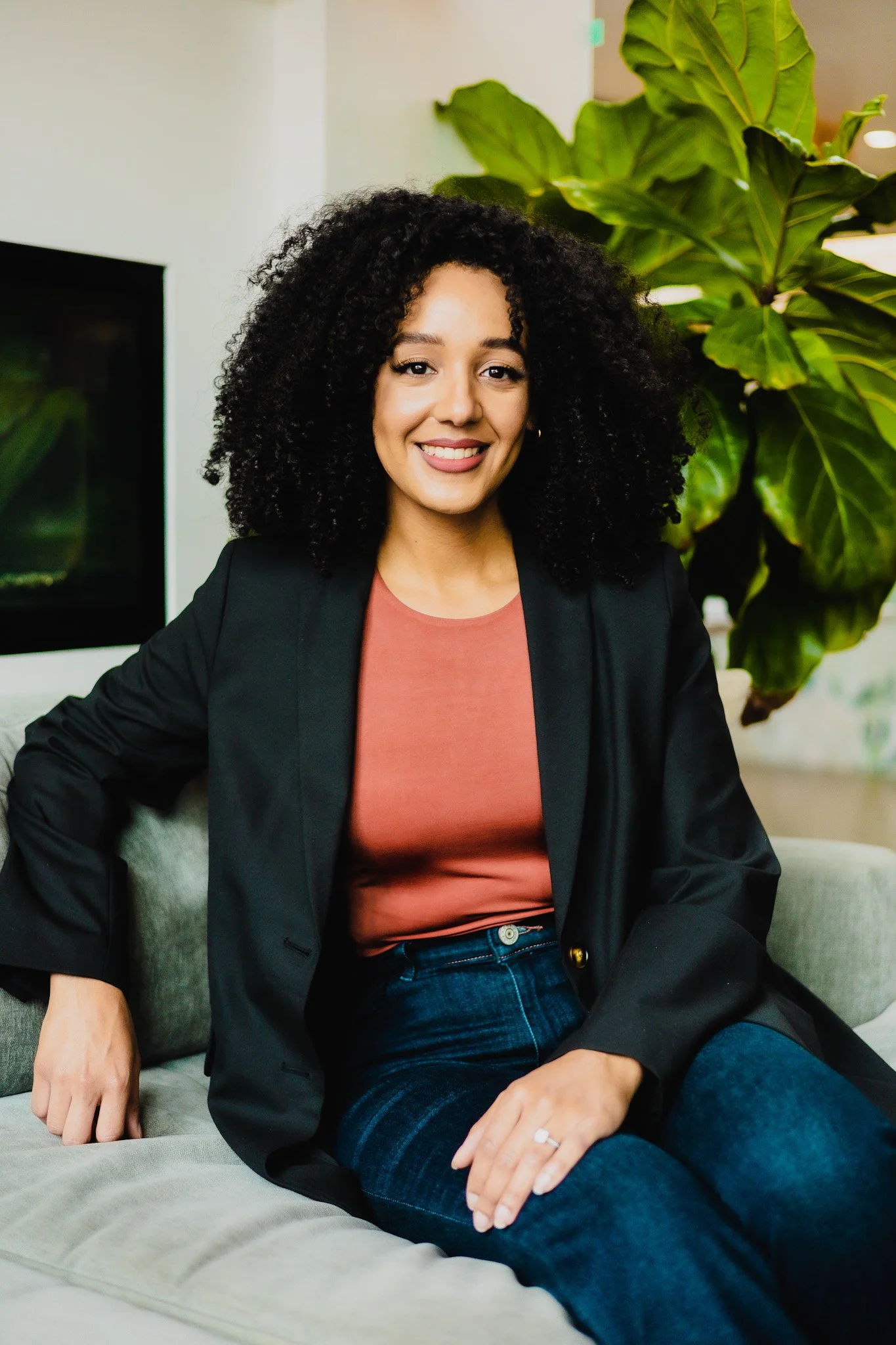 A woman with curly black hair smiling, sitting on a beige couch in a room with a large green houseplant behind her, wearing a black blazer, rust-colored top, and blue jeans.