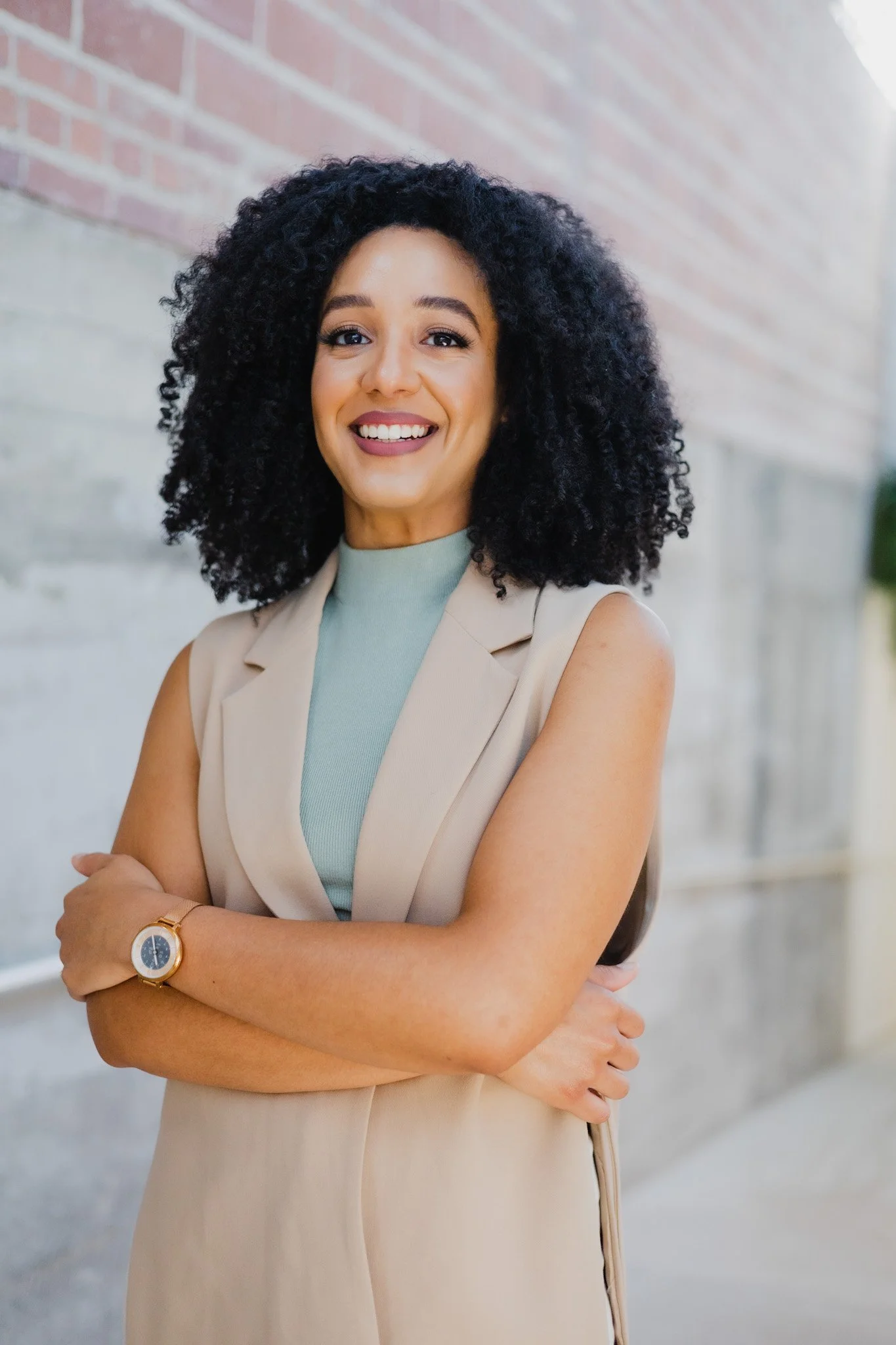 A smiling woman with curly black hair, wearing a beige sleeveless blazer over a light blue top, standing outdoors in front of a brick wall, with her arms crossed and wearing a gold watch.