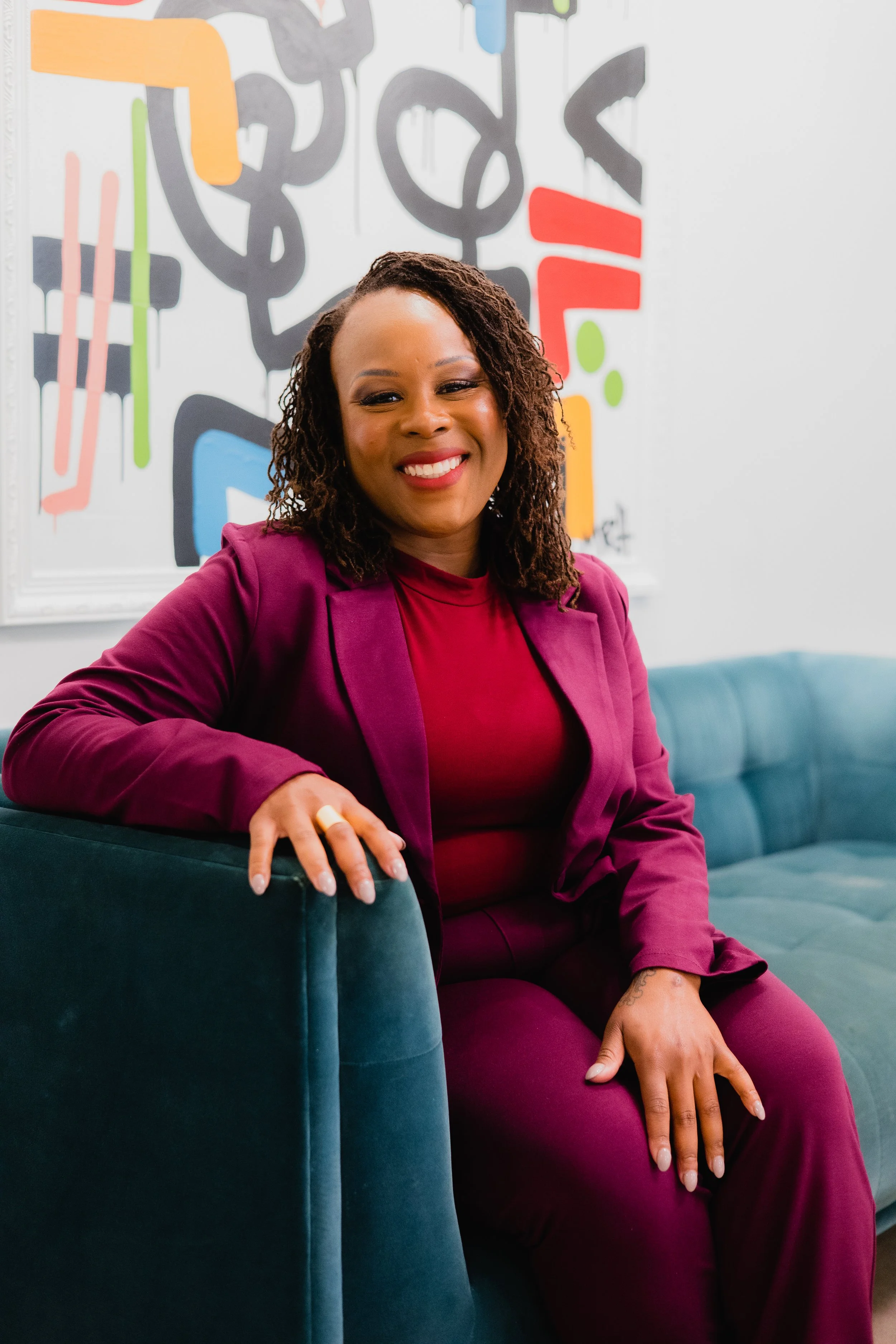 A woman with shoulder-length curly hair, wearing a magenta blazer and matching pants, sitting on a teal velvet sofa. She is smiling, with her left hand resting on the sofa arm and her right hand on her knee. Behind her is a colorful abstract wall art