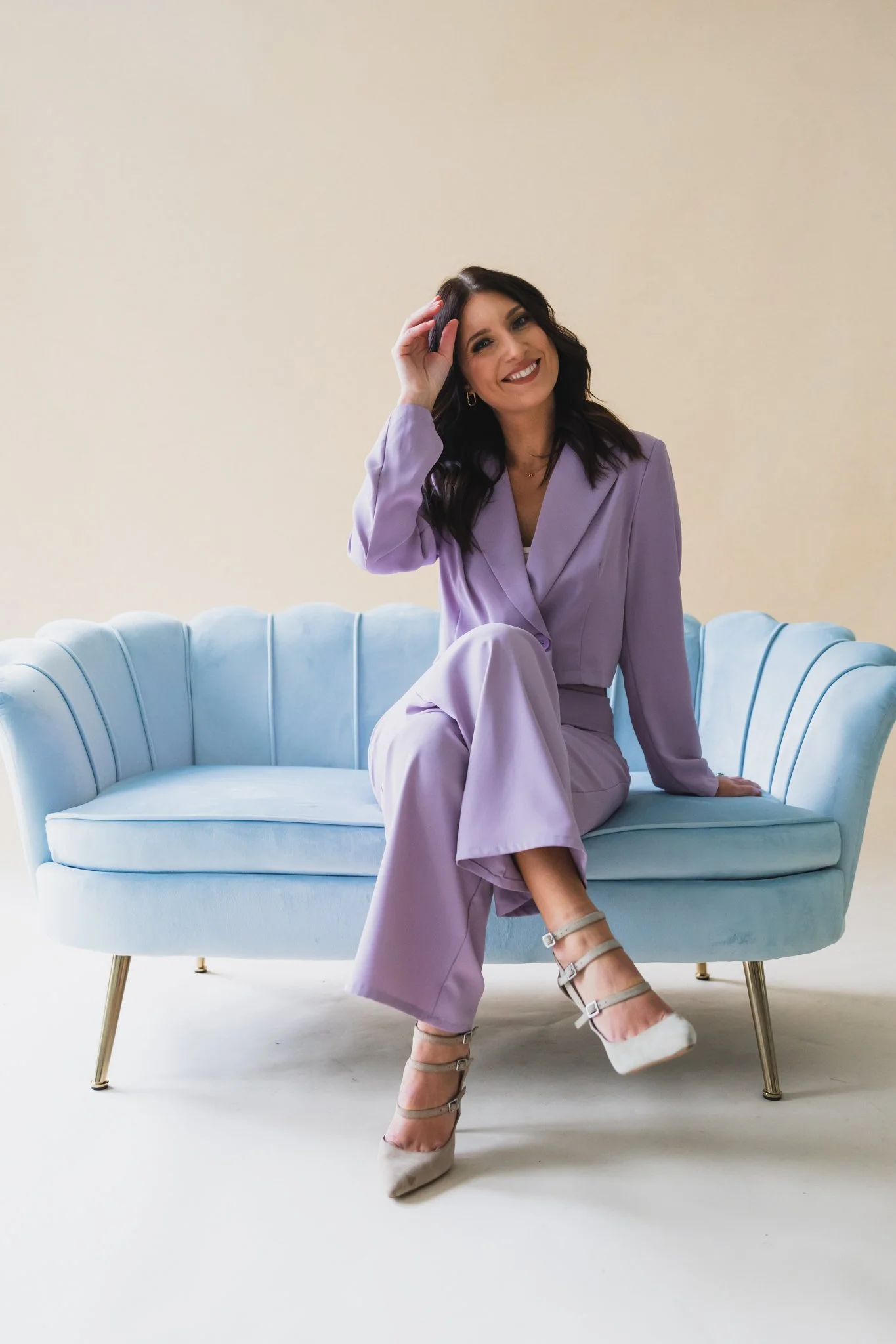 A woman sitting on a light blue vintage sofa, wearing a lavender pantsuit with white high-heeled shoes, smiling and adjusting her hair against a plain cream-colored background.