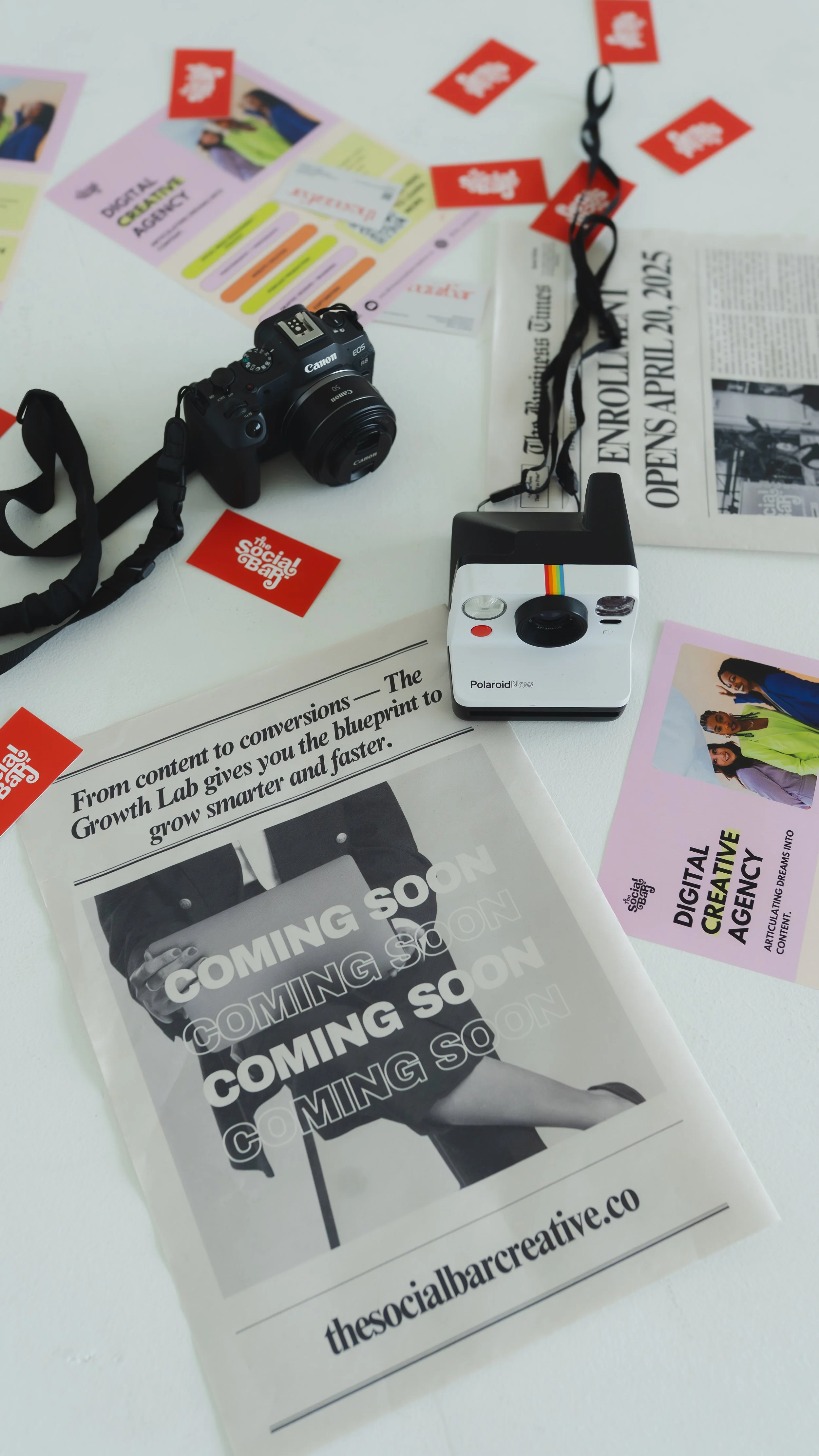 A white table with various items including a Canon camera, a Polaroid instant camera, a printed 'Coming Soon' flyer, a newspaper announcing an open event, some colorful flyers, and red stickers with white text reading 'The Social Bar'.