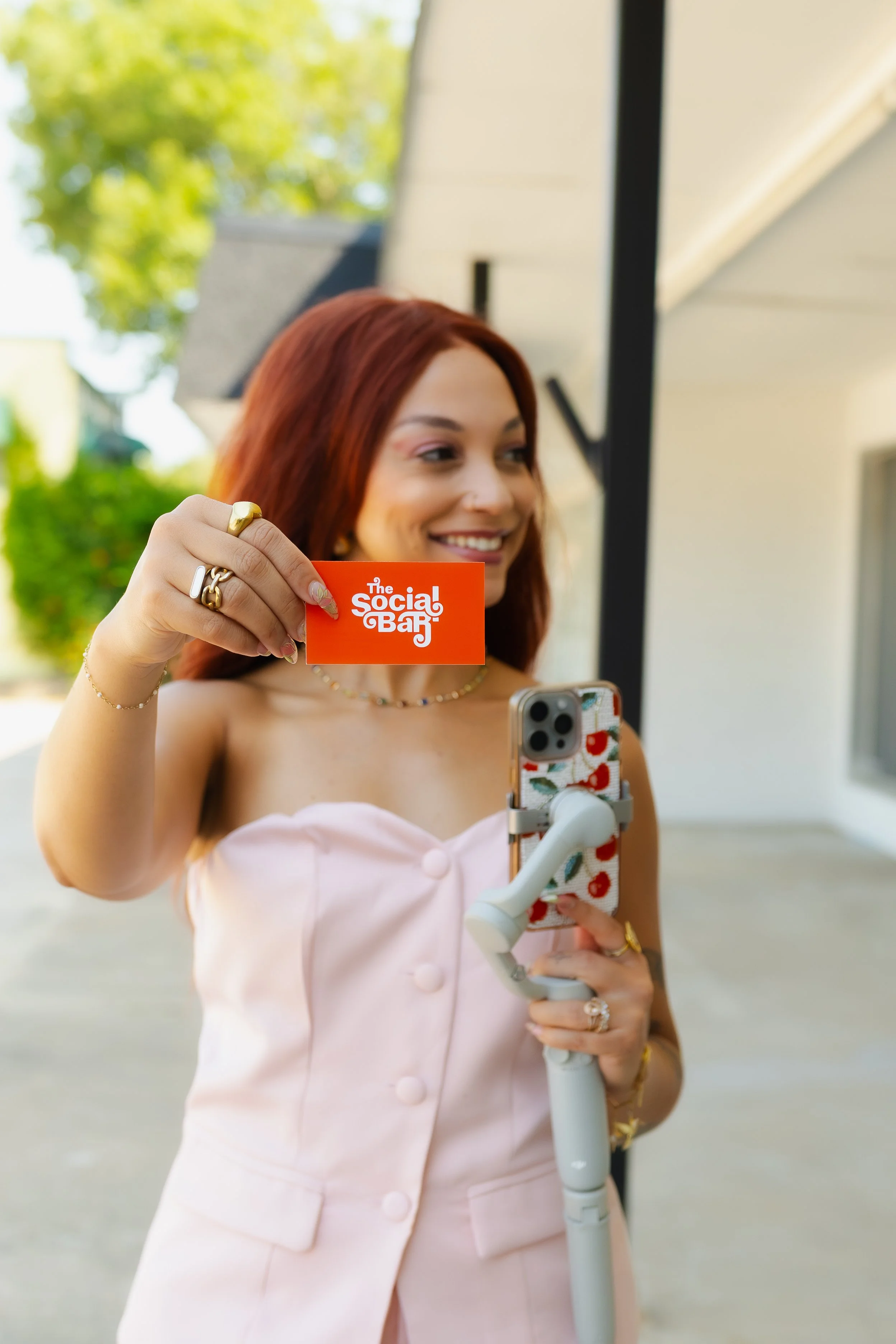 A woman with red hair smiles and holds a bright orange card that says 'The Social Bar'. She stands outside, wearing a light pink dress with buttons, and is using a white electric scooter. Behind her, there are trees and a building.
