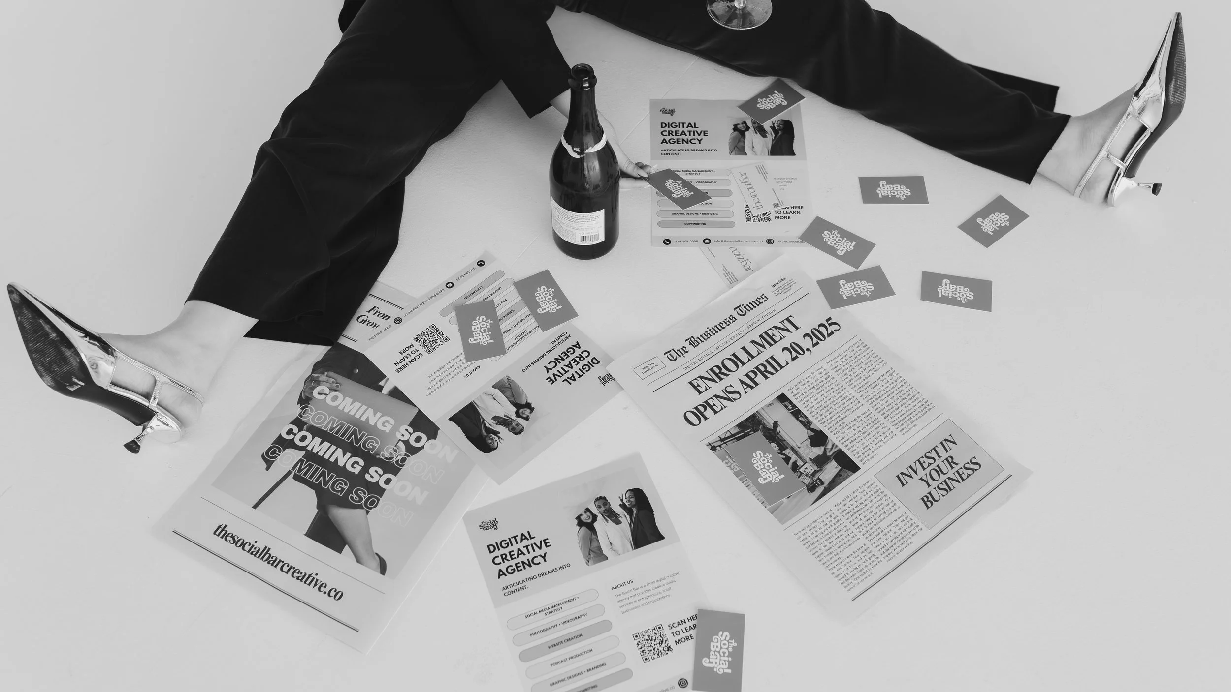 A person wearing shiny silver high heels and black clothing sitting with legs spread on a white floor surrounded by various flyers, newspapers, a bottle, and business cards related to a digital creative agency.