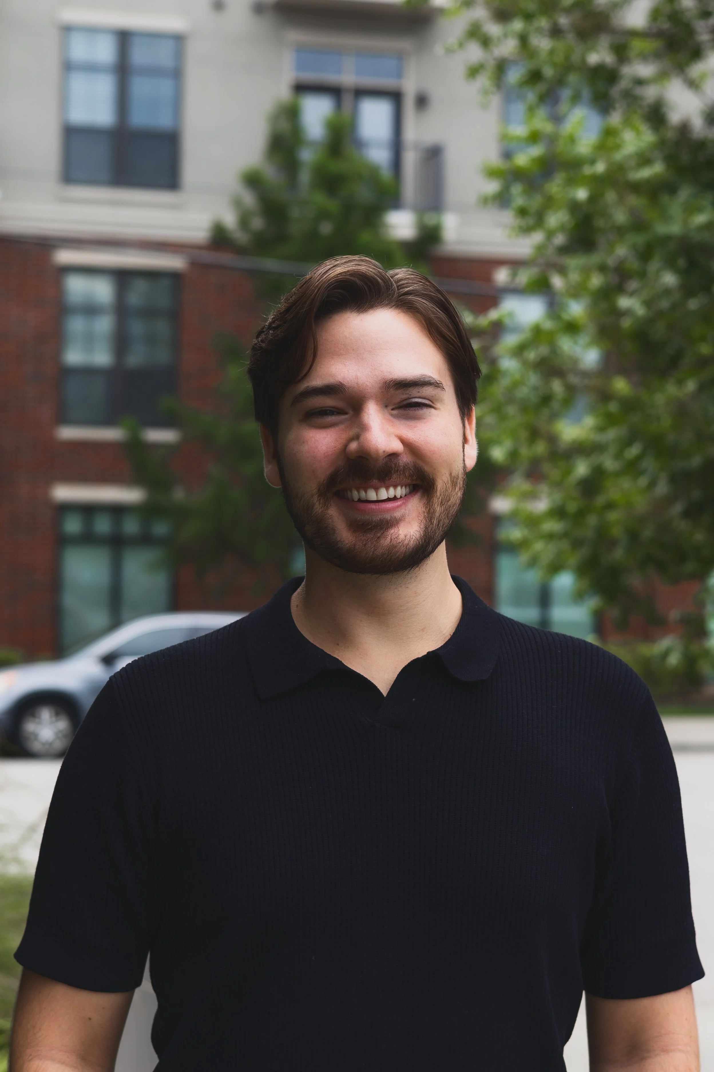 Close-up of a smiling young man with dark hair and a beard, wearing a black polo shirt, outdoors with trees and residential buildings in the background.