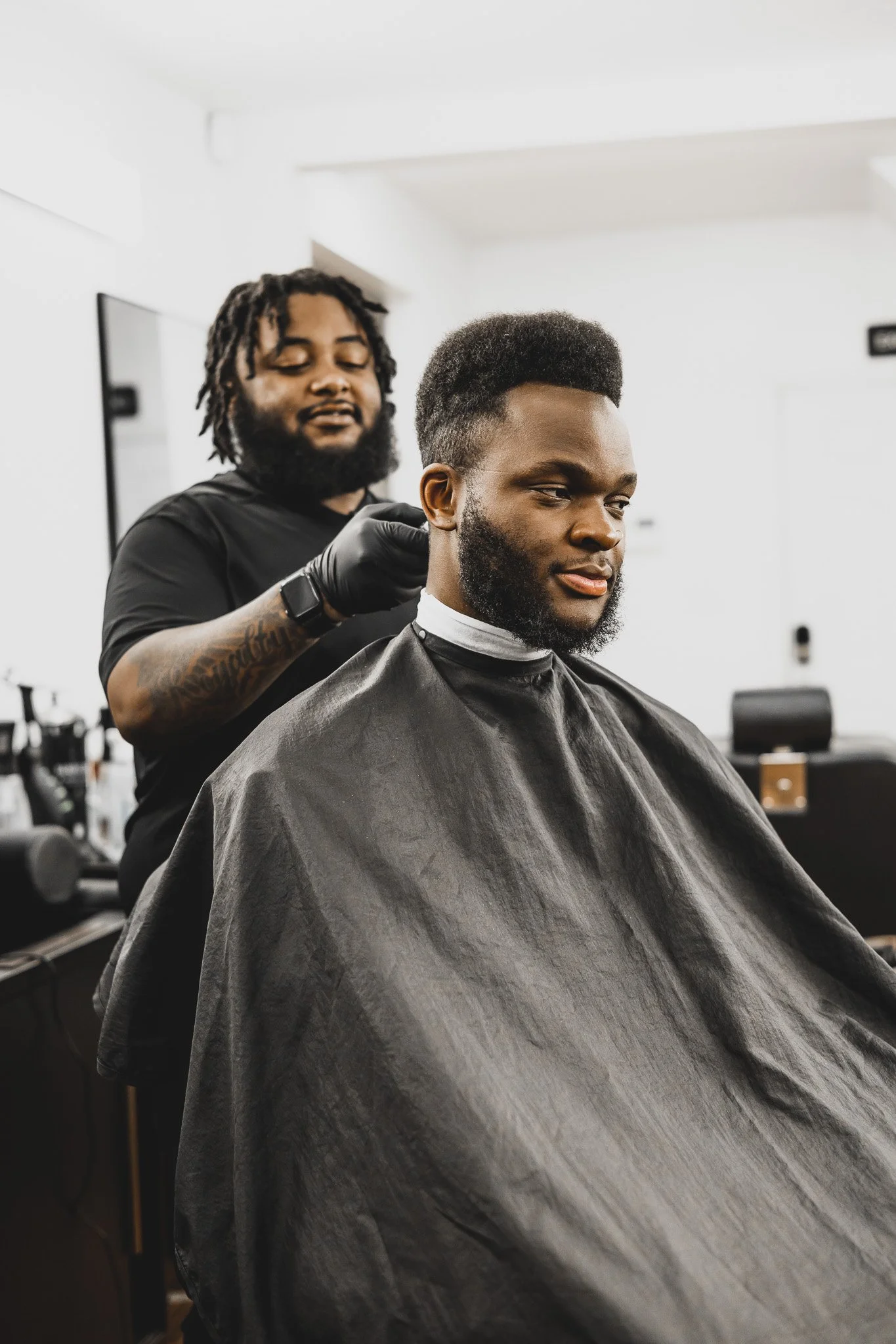 A man with a beard sitting in a barber's chair. A barber with dreadlocks and tattoos is trimming his hair. The setting appears to be a modern barbershop.