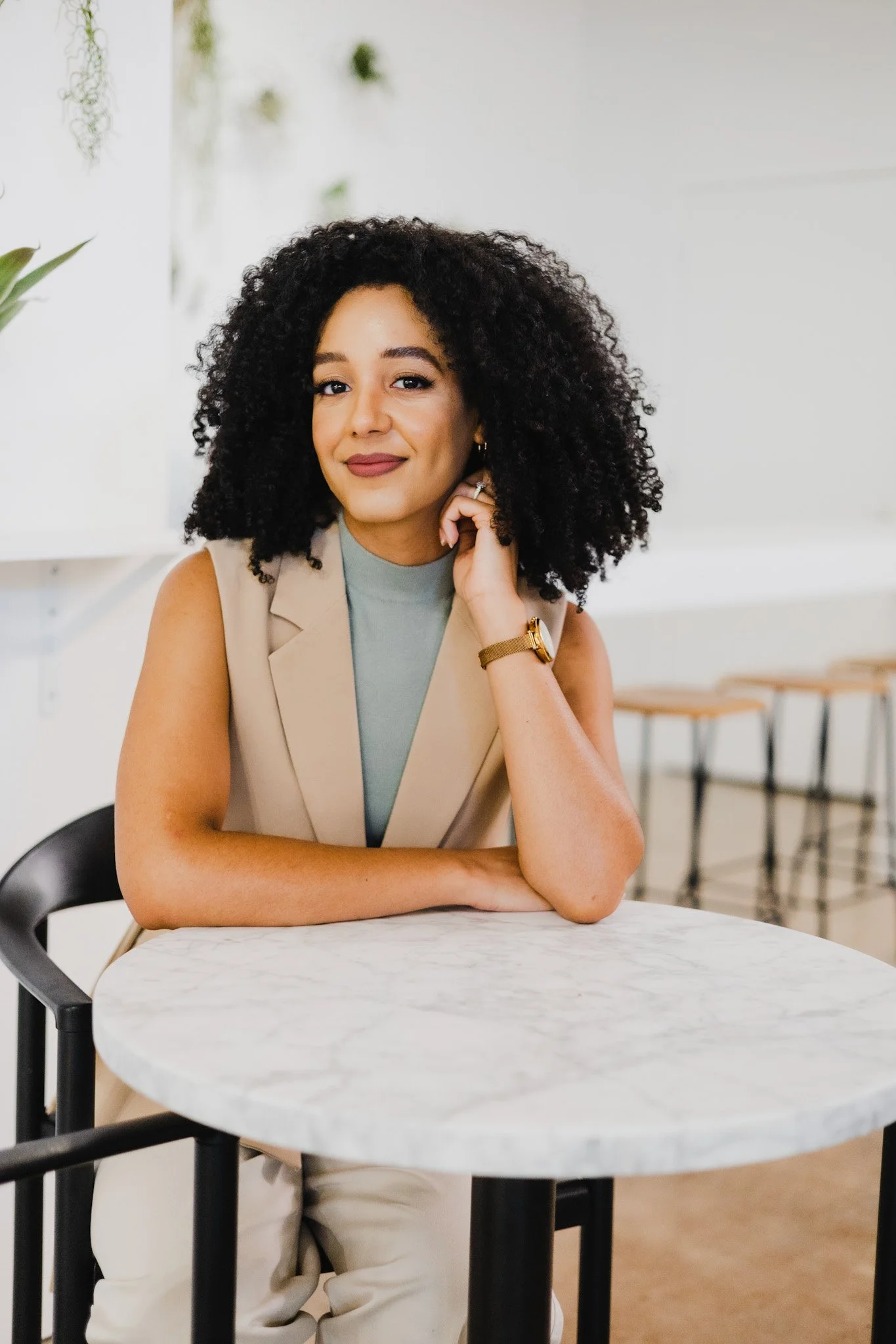 A woman with curly black hair, wearing a grey sleeveless top and beige vest, sitting at a round marble table in a bright indoor space, resting her chin on her hand, smiling at the camera.