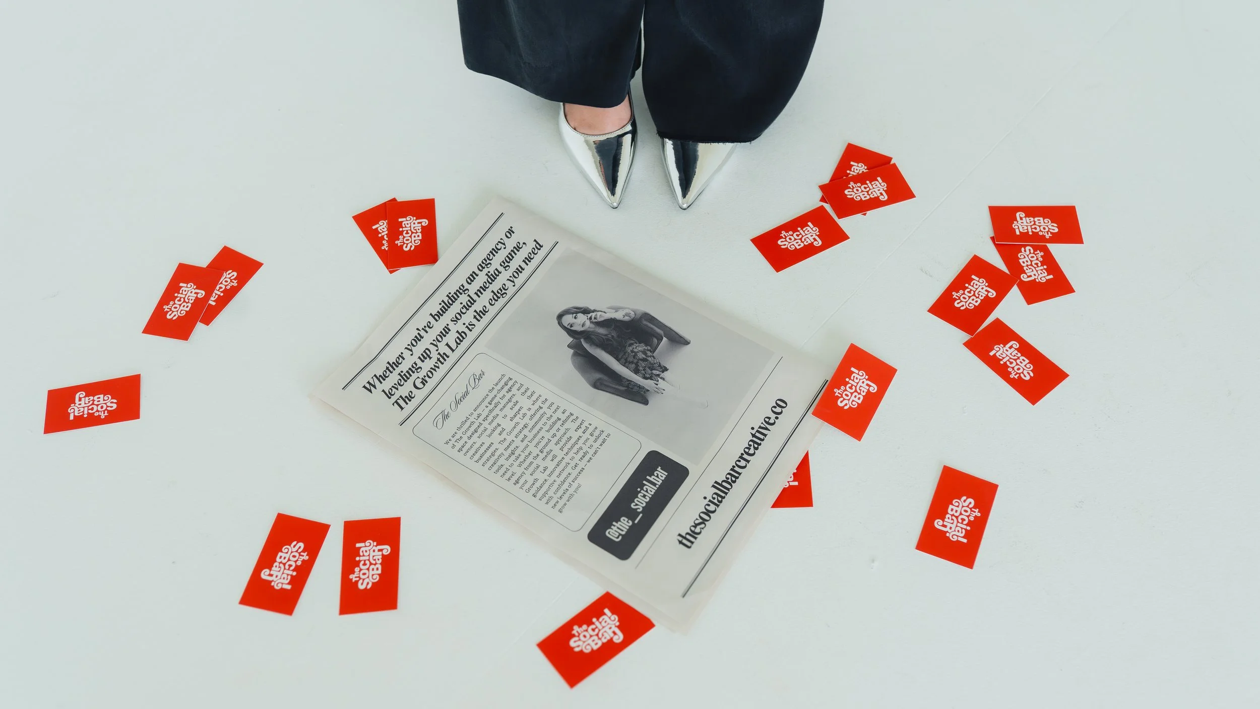 A person's feet wearing silver high heels standing on a white floor, surrounded by scattered red stickers that have white text reading 'The Social Gab.' Part of a paper with a black-and-white photo of a woman and text about building an agency and soc