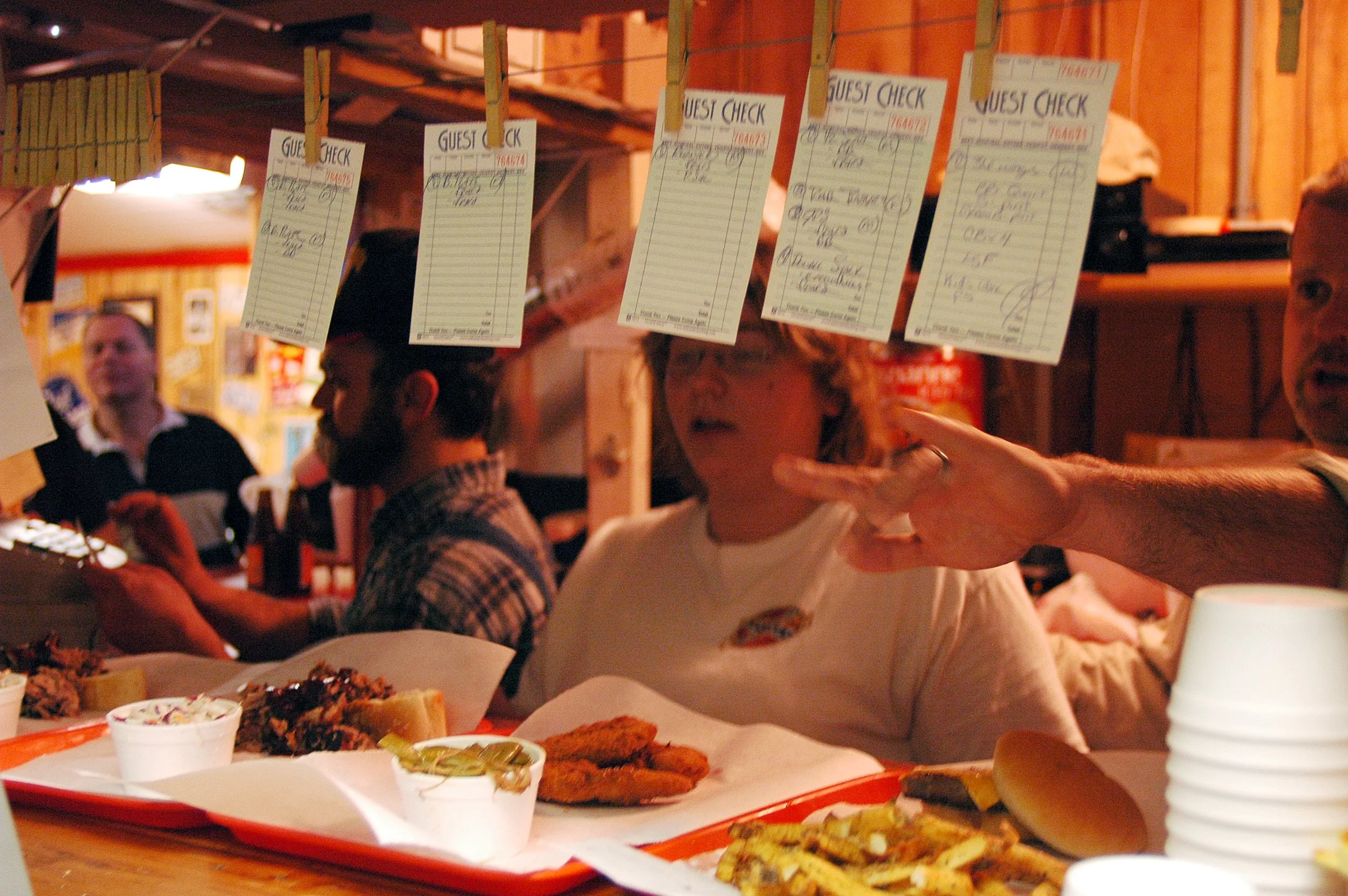 Busy restaurant with diners eating barbecue, trays of food, and handwritten guest checks hanging above the tables.