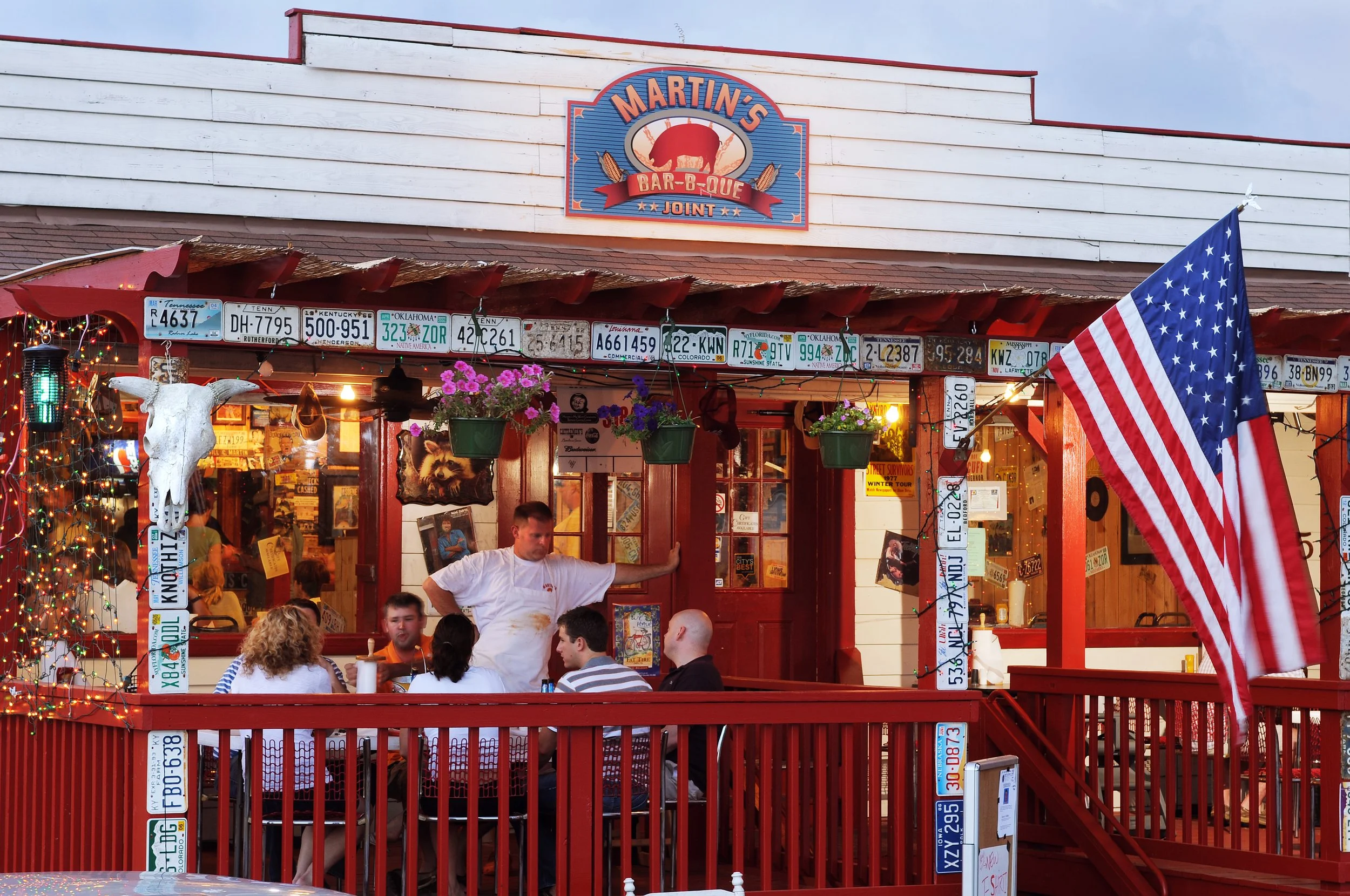 Martin's Bar-B-Que Joint restaurant exterior with vintage license plates, patio diners, hanging flowers, string lights, and an American flag