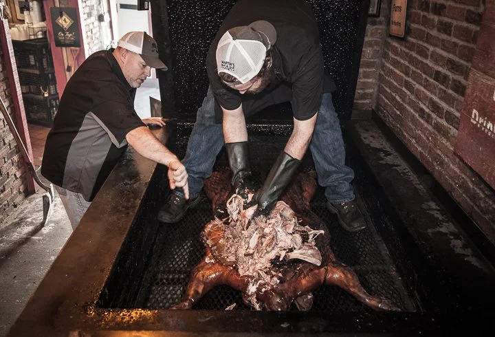 Two workers in protective gloves prepare a roasted whole pig in a commercial barbecue pit.