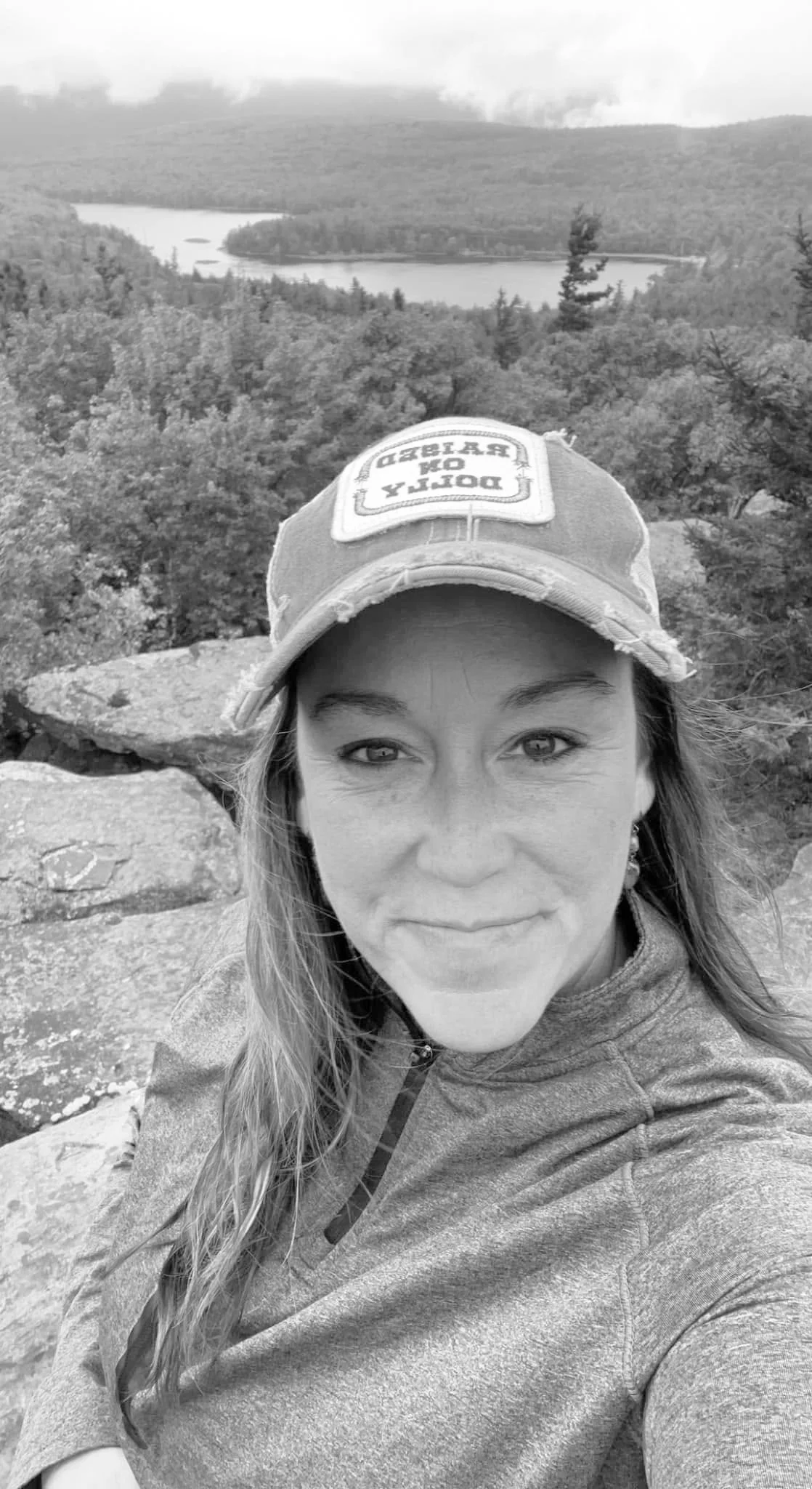A smiling woman with long hair and a ball cap on with a scenic lake backdrop