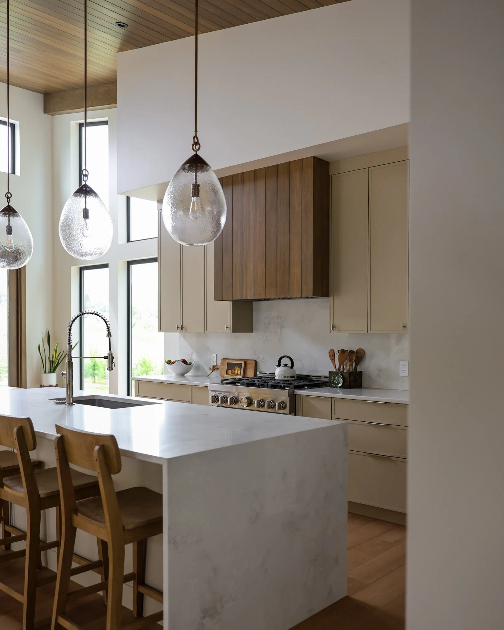 Interior view of a modern kitchen with a white island and wooden dining chairs, pendant lights hanging from a wooden ceiling, large windows letting in natural light, and a stove with a kettle on top.