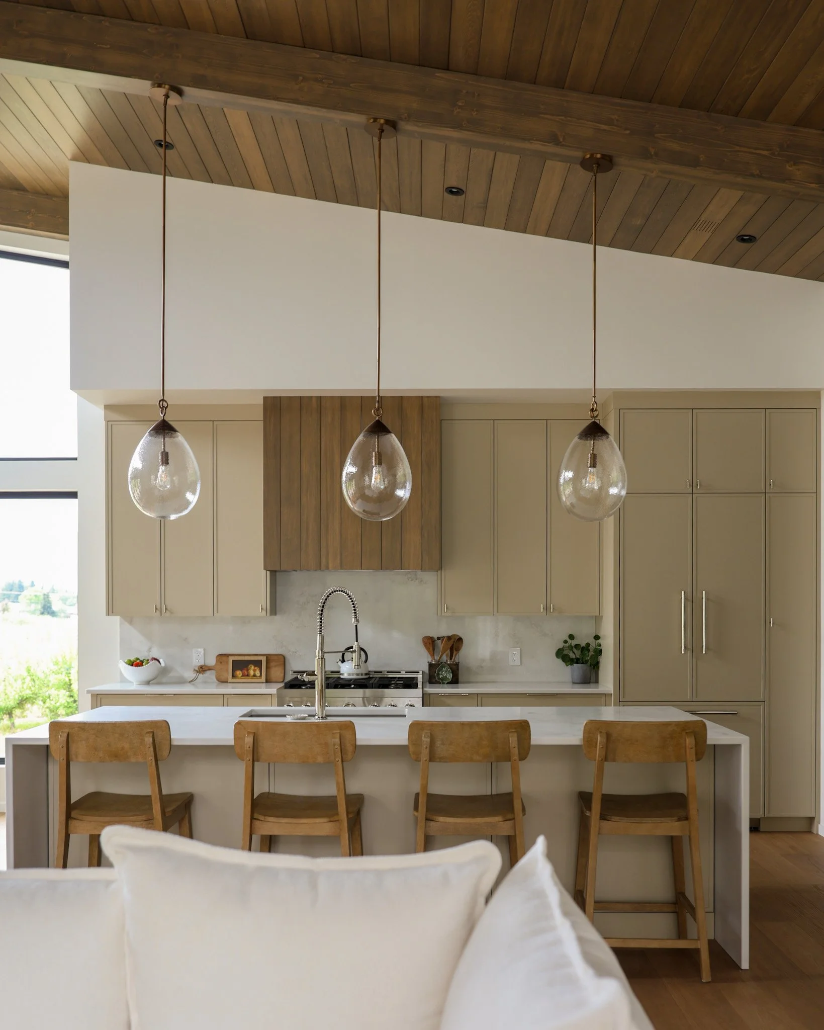 View of a modern kitchen with a white island counter, wooden bar stools, beige cabinetry, a stainless steel stove, and hanging glass pendant lights. The ceiling is made of wood panels and there is a window with a view of greenery outside.