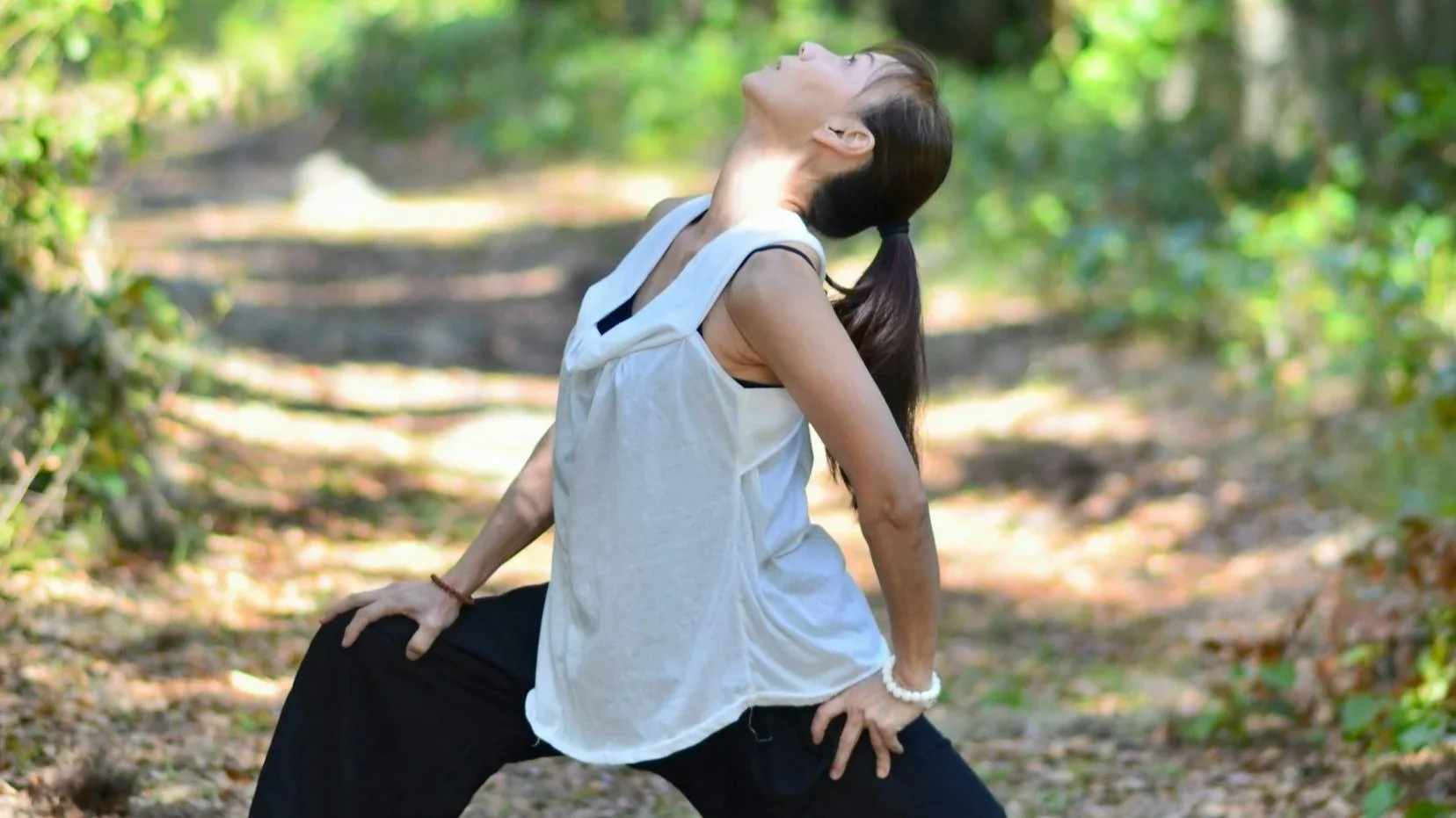 Woman in a white sleeveless hoodie and black pants practicing yoga or stretching outdoors on a forest trail.