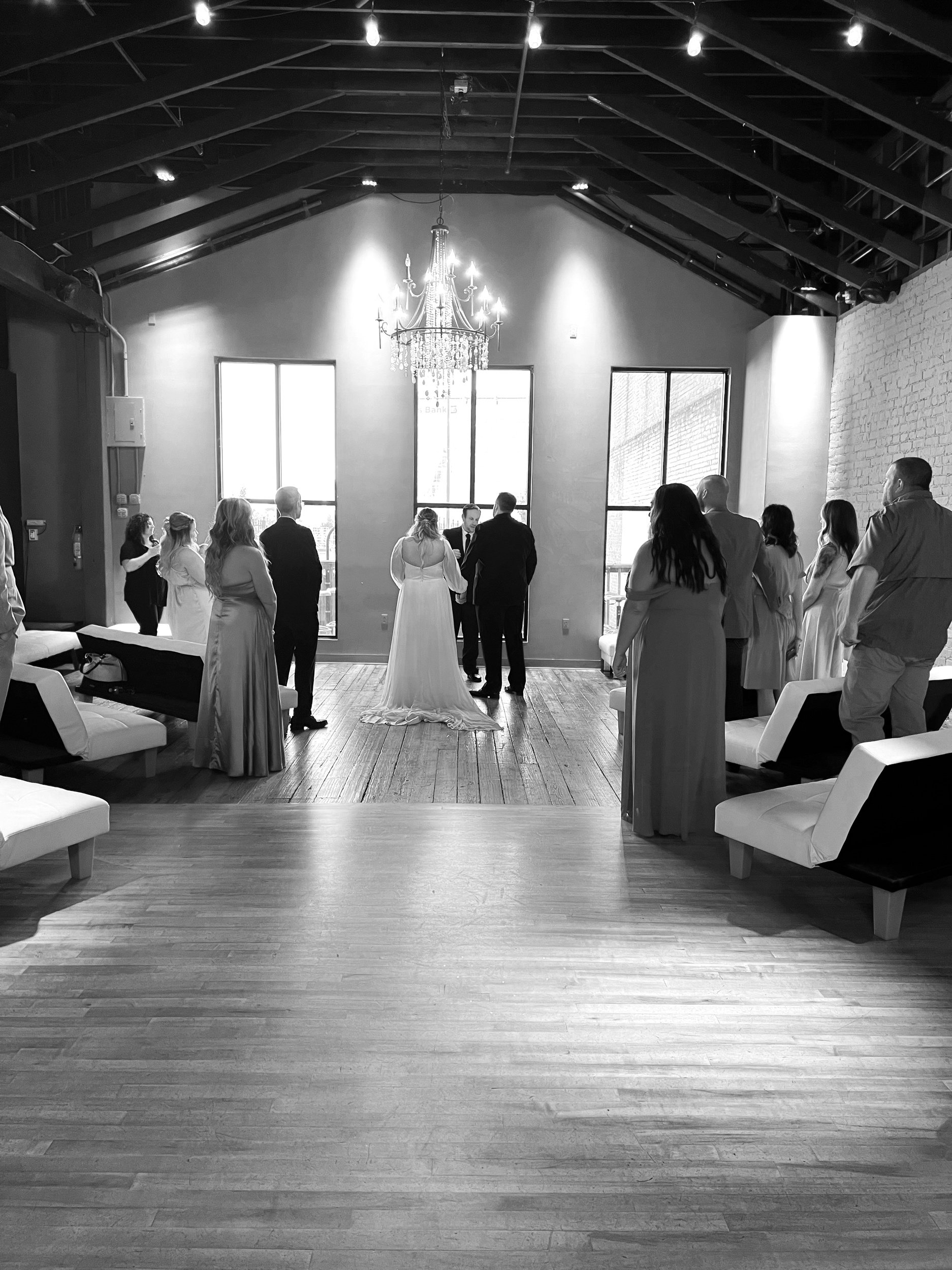 Black and white image of a wedding ceremony with a couple standing together in front of a group of people in a room with a high ceiling and chandelier.