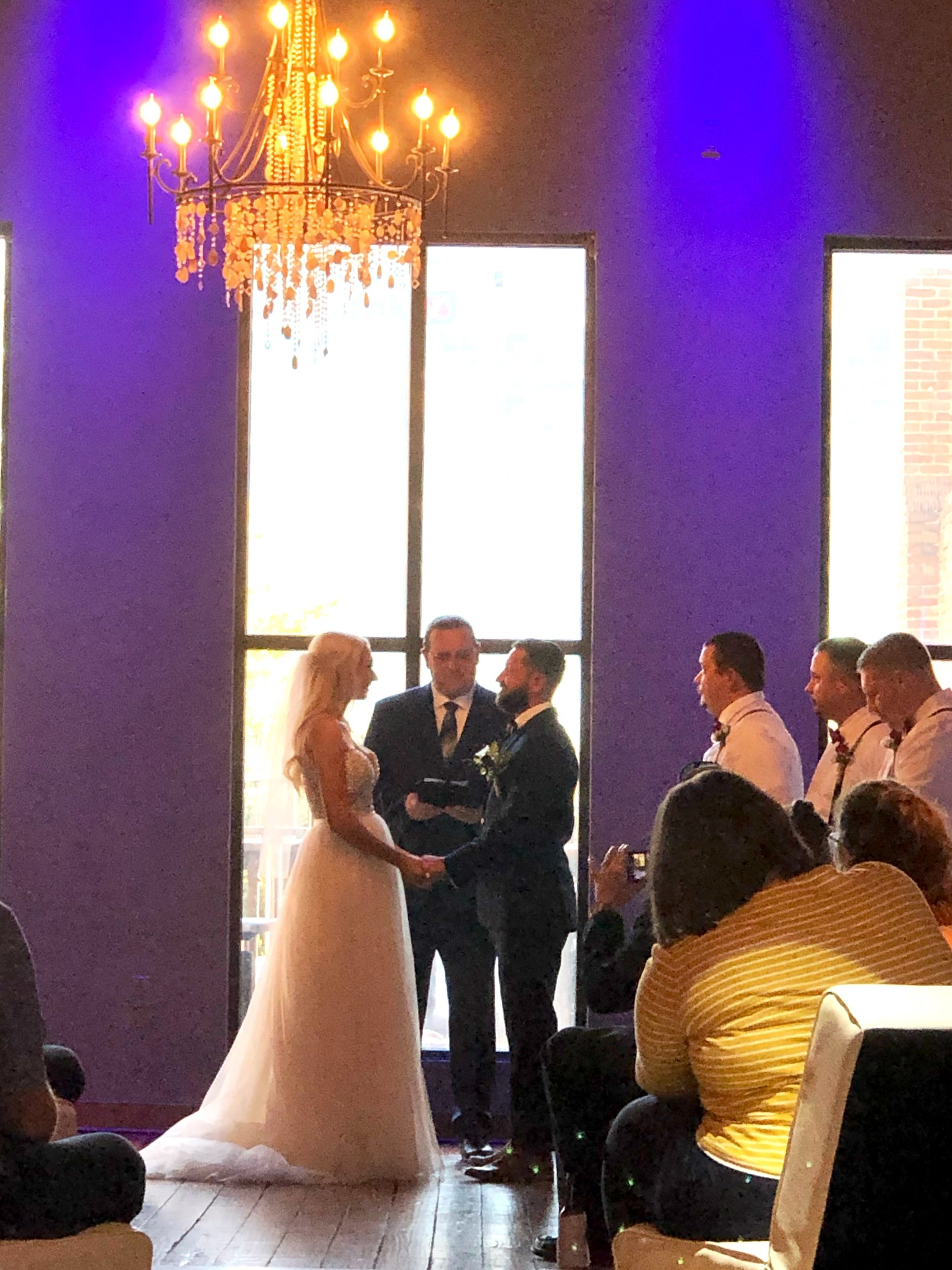 Wedding ceremony with a couple exchanging vows under a chandelier, guests seated and a wedding party present.