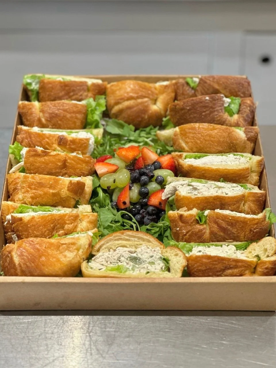 A tray of assorted sandwich croissants filled with lettuce and other ingredients, topped with a variety of fresh berries and grapes in the center.