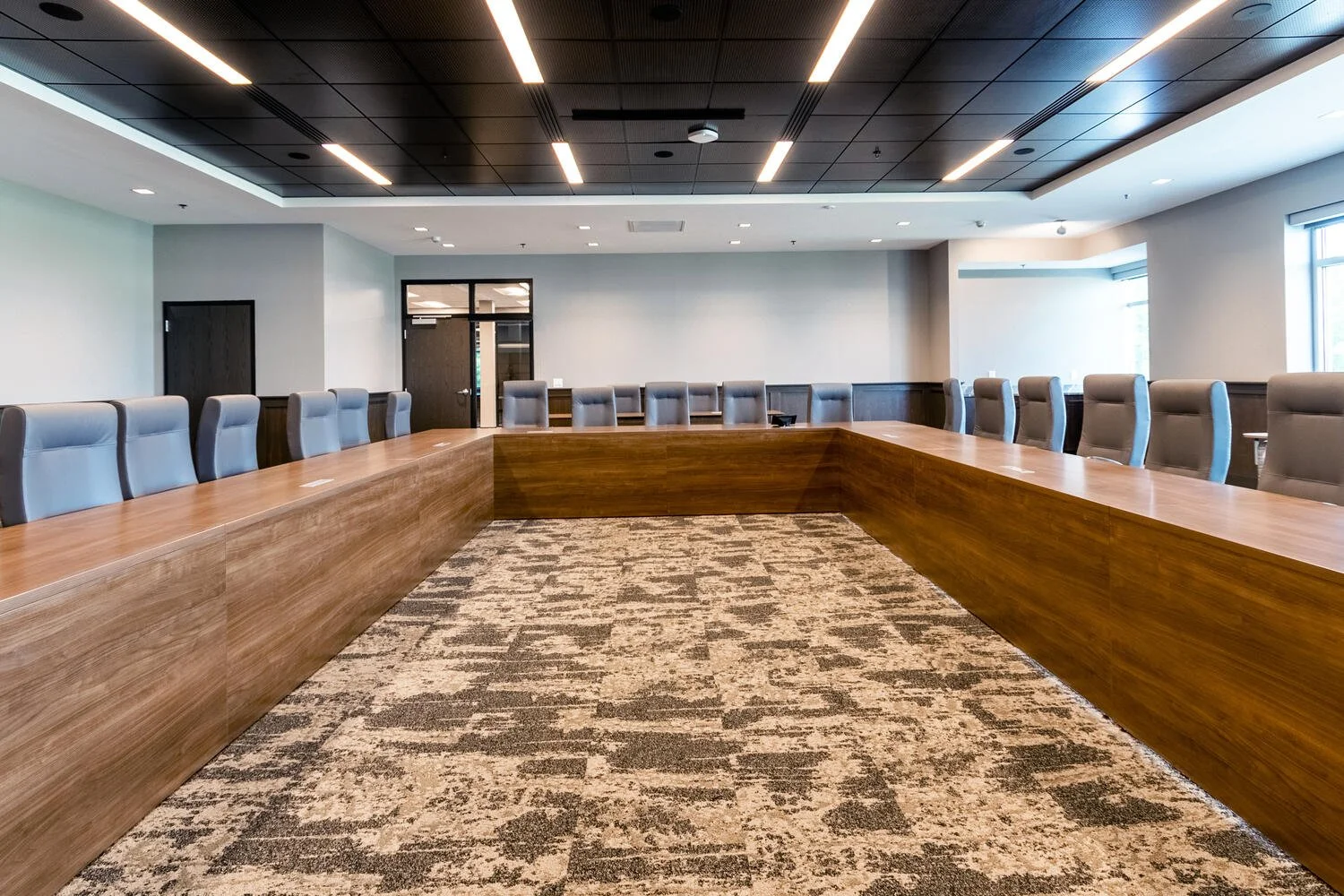 Empty conference room with a large U-shaped wooden table surrounded by gray chairs, carpeted floor, and large windows letting in natural light.