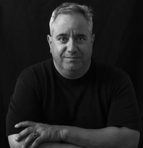 Black and white portrait of a smiling man with short hair, wearing a dark long-sleeve shirt, with arms crossed, against a dark background.