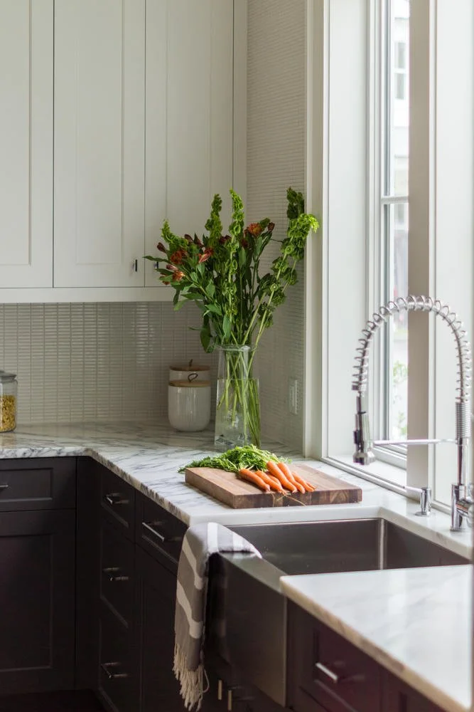 Kitchen with white upper cabinets, dark lower cabinets, a marble countertop, a sink, a large window, a glass vase with greenery and flowers, a cutting board with carrots, and a striped dish towel.