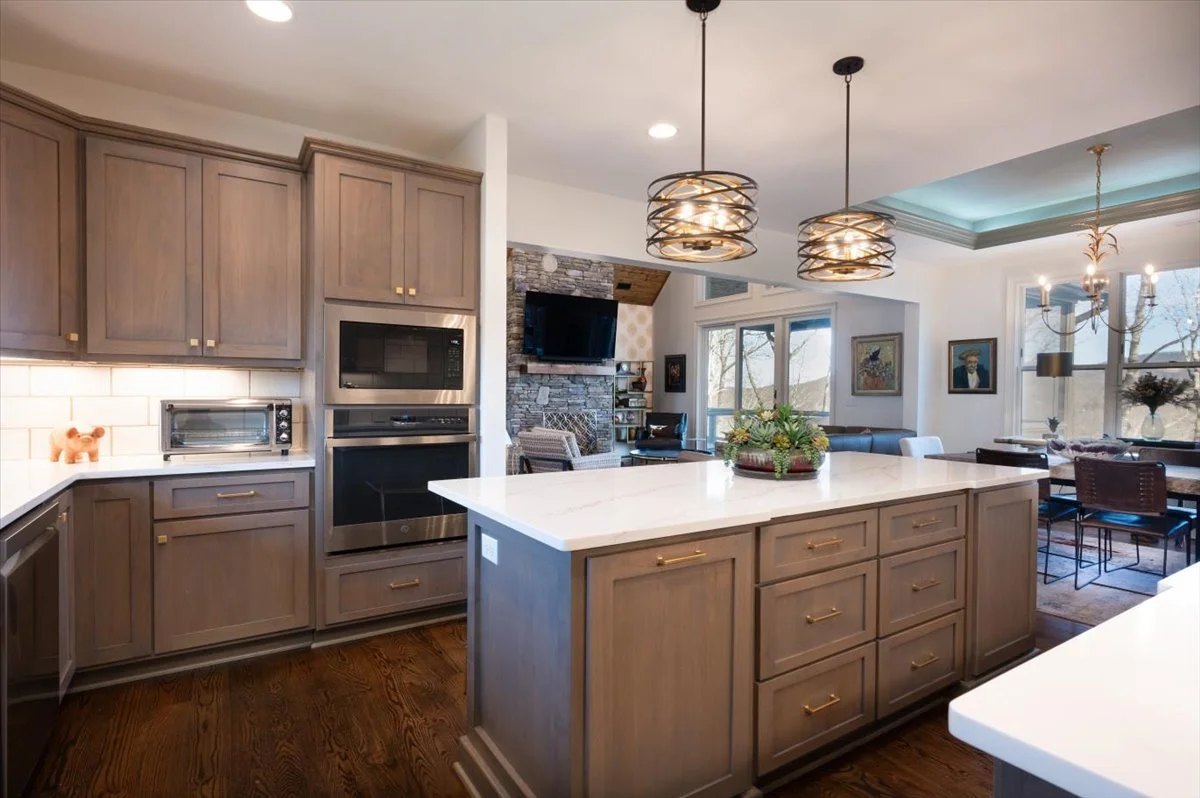 Modern kitchen with wood cabinets, white countertops, and a large island in the center. The background features a living area with a TV, stone wall, and dining space with a chandelier. Pendant lights hang above the island, and sunlight streams through large windows.