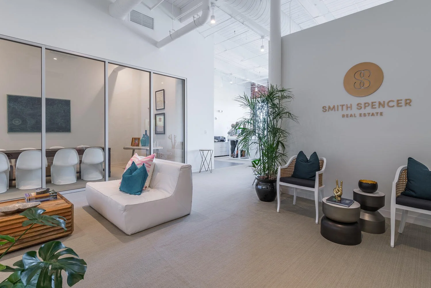 Interior of a modern real estate office with white walls, a sign reading "Smith Spencer Real Estate," seating area with white and black chairs, a white sofa with colorful pillows, a large potted plant, and a glass-enclosed conference room.