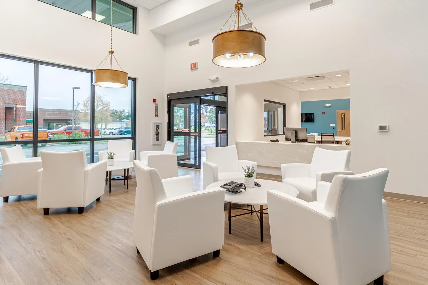 Lobby area with white armchairs arranged around small round tables, large windows, and a reception desk, with a view of parked cars outside.