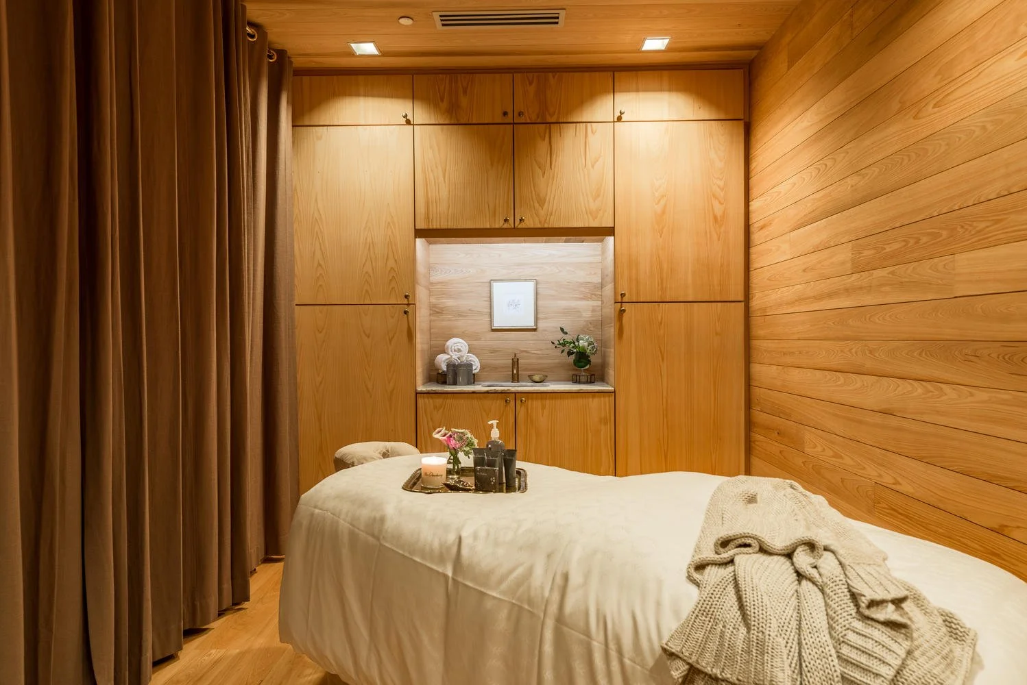 A cozy massage room with wooden walls and ceiling, a massage table with a beige blanket, a tray with flowers and spa products, a small cabinet area with towels, a framed picture, and decorative items, partially covered by curtains.